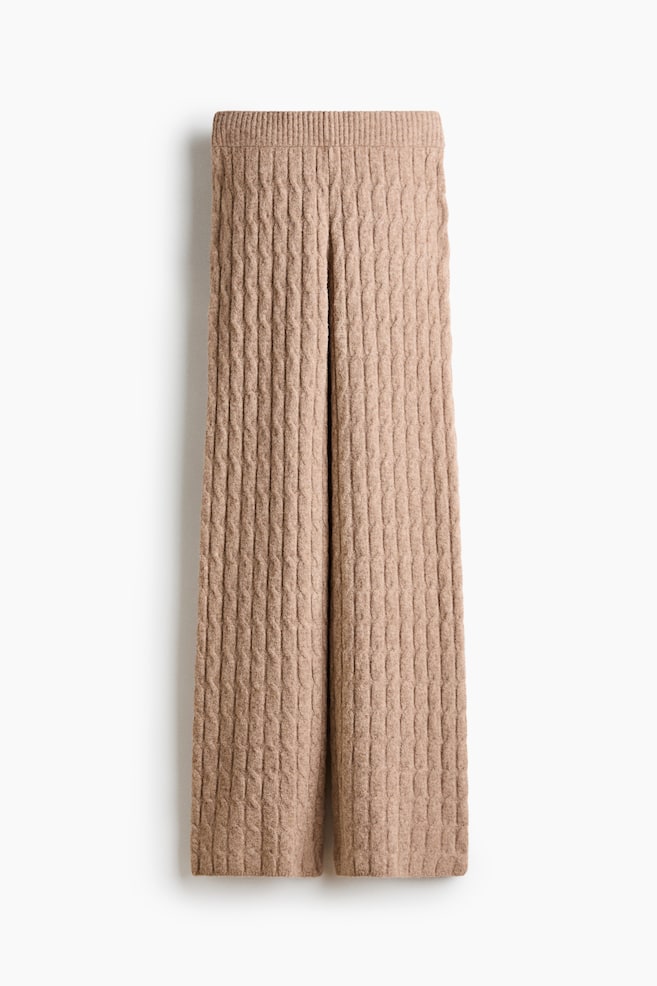 Diese hellbraunen Strickhosen hängen in einer frontalen Ansicht und präsentieren eine weite Silhouette mit einem durchgehenden strukturierten Zopfmuster sowie einem breiten, gerippten Bund. - 1