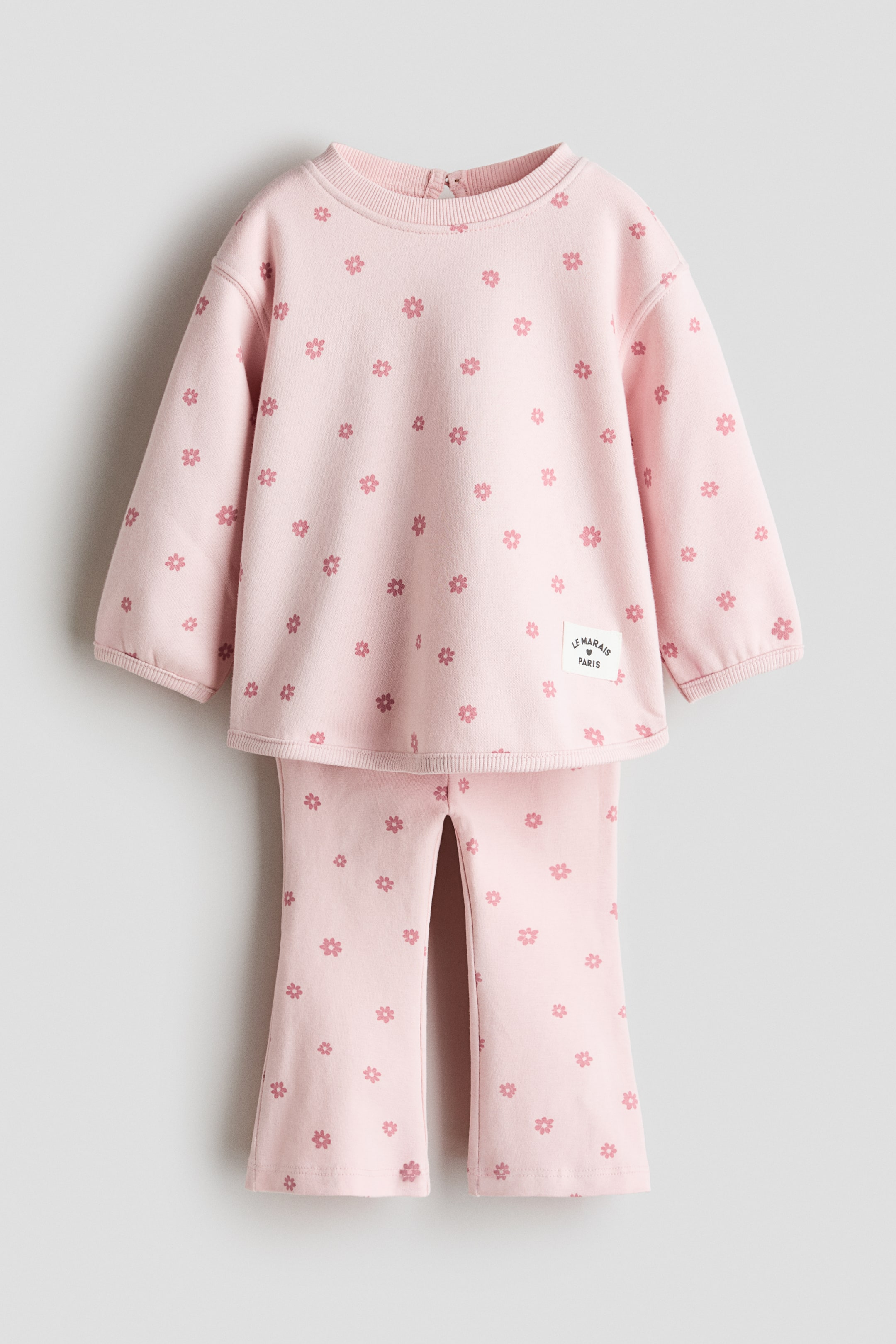 View larger image: A pink two-piece children's set is laid out flat, showcasing a coordinated top and pants, both adorned with an all-over pattern of small pink flowers. The crewneck top features a ribbed collar, long sleeves with ribbed cuffs, a ribbed hem, a small zipper closure at the back of the neck, and a rectangular "LE MARAIS PARIS" label on the lower right front. The pants feature flared legs with small slits at the front bottom hem.
