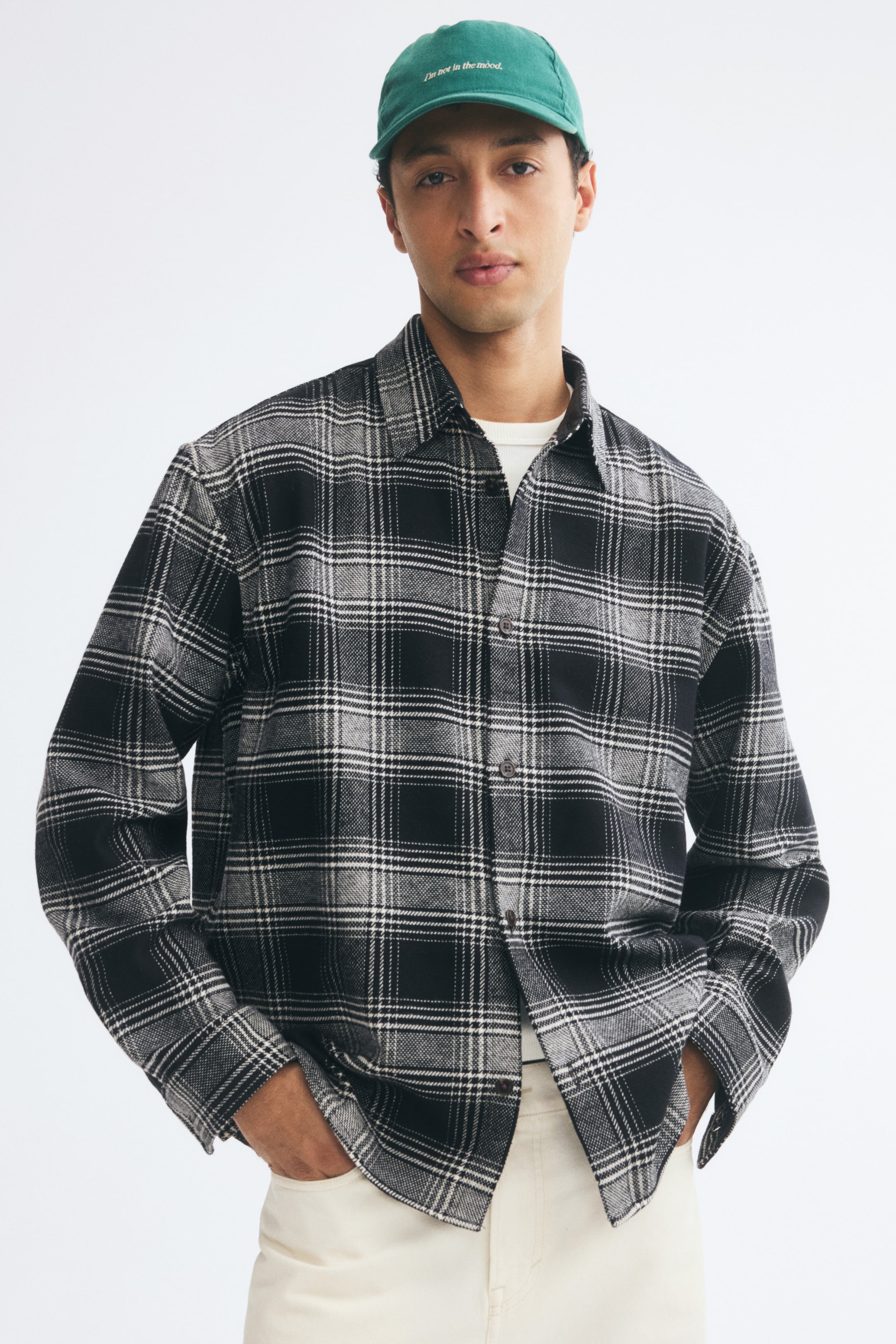 Agrandir l'image: Une surchemise à carreaux en tissu texturé noir, gris et blanc est portée par le mannequin par-dessus un T-shirt blanc et un pantalon clair. La surchemise présente un col, une fermeture boutonnée sur le devant et des manches longues, tandis que le mannequin est debout, les mains dans les poches.