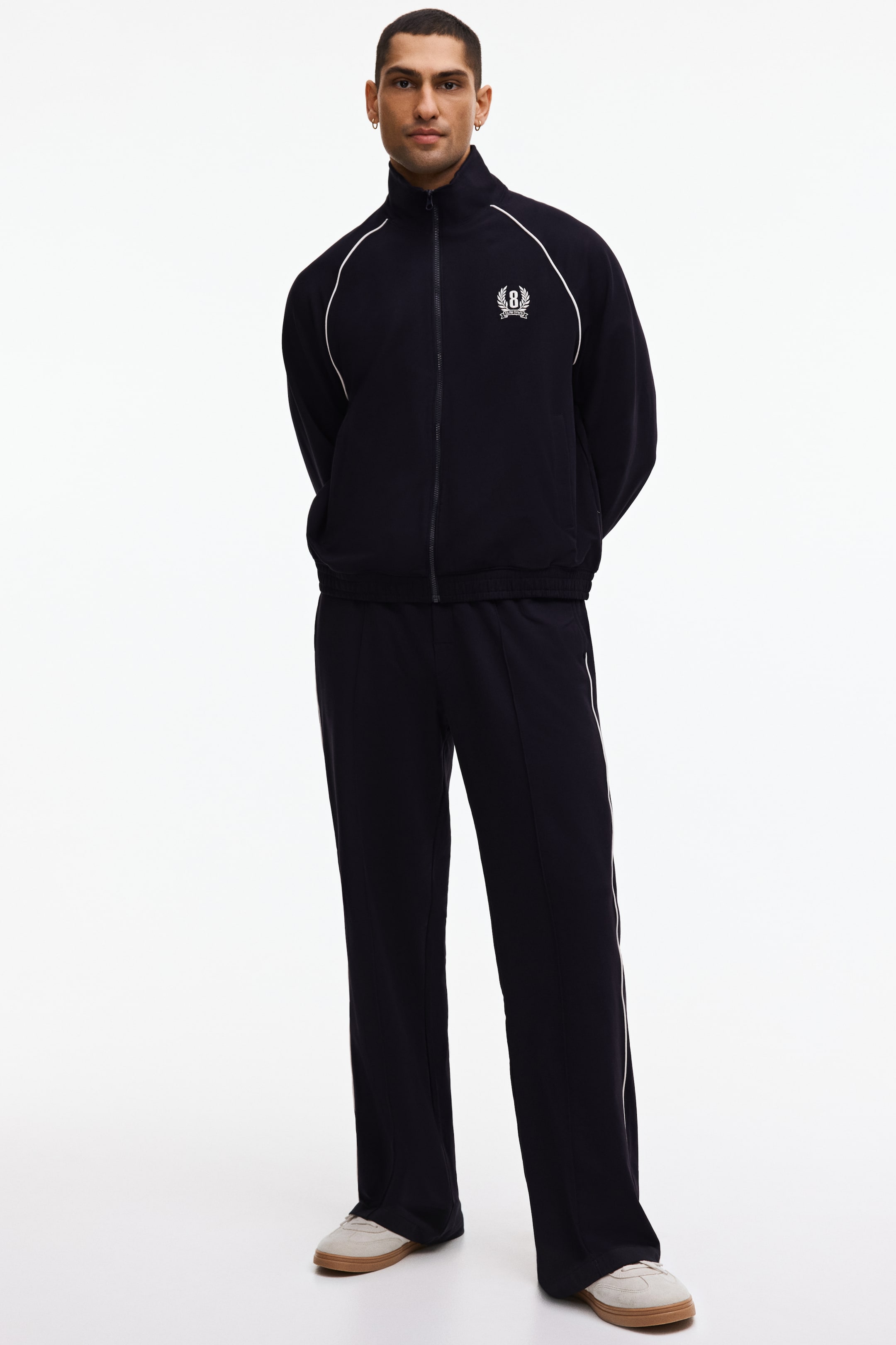 查看大圖: 這是一套海軍藍運動服，包含一件全拉鍊的立領外套，肩袖上有白色滾邊，左胸繡有徽章標誌。配套的直筒褲在側縫和每條褲腿的前面中縫都有白色滾邊。