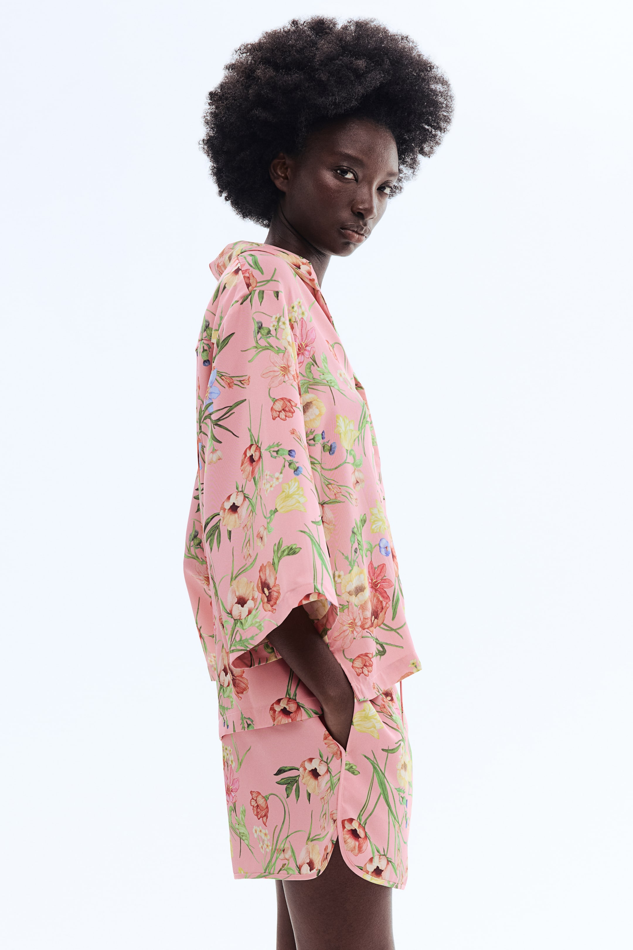 View larger image: A pink floral pajama set is showcased from a slight side angle, featuring a loose-fitting short-sleeve shirt and matching shorts. The shirt is styled with a revere collar and an all-over print of vibrant flowers and green leaves, while the shorts display a curved hem and visible pockets with hands resting inside.