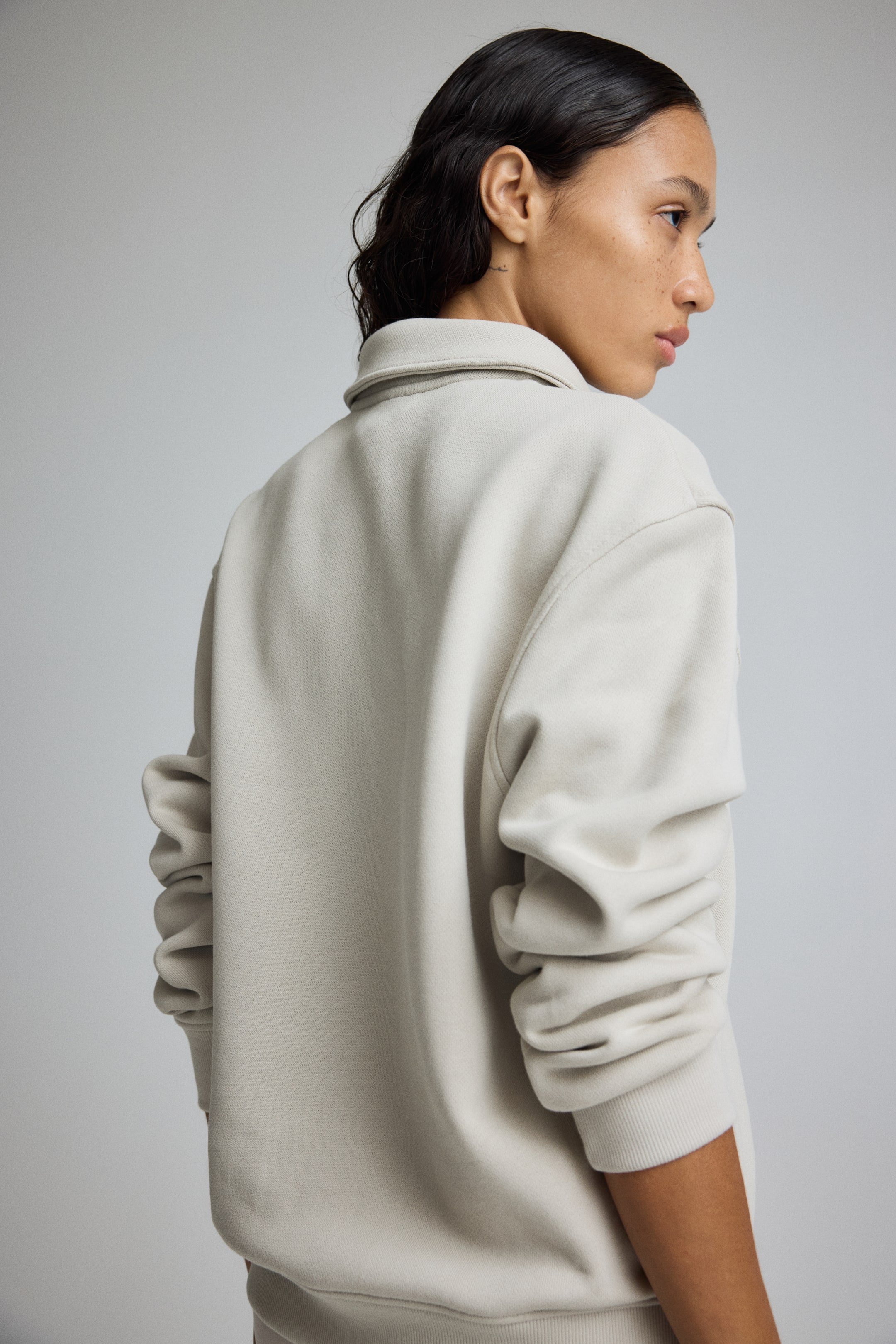 Grotere afbeelding bekijken: Een lichtbeige sweatshirt met kraag wordt gepresenteerd vanaf de achterkant en de rechterkant. Het heeft een gestructureerde kraag, met lange, ontspannen mouwen die toelopen naar geribbelde boorden.