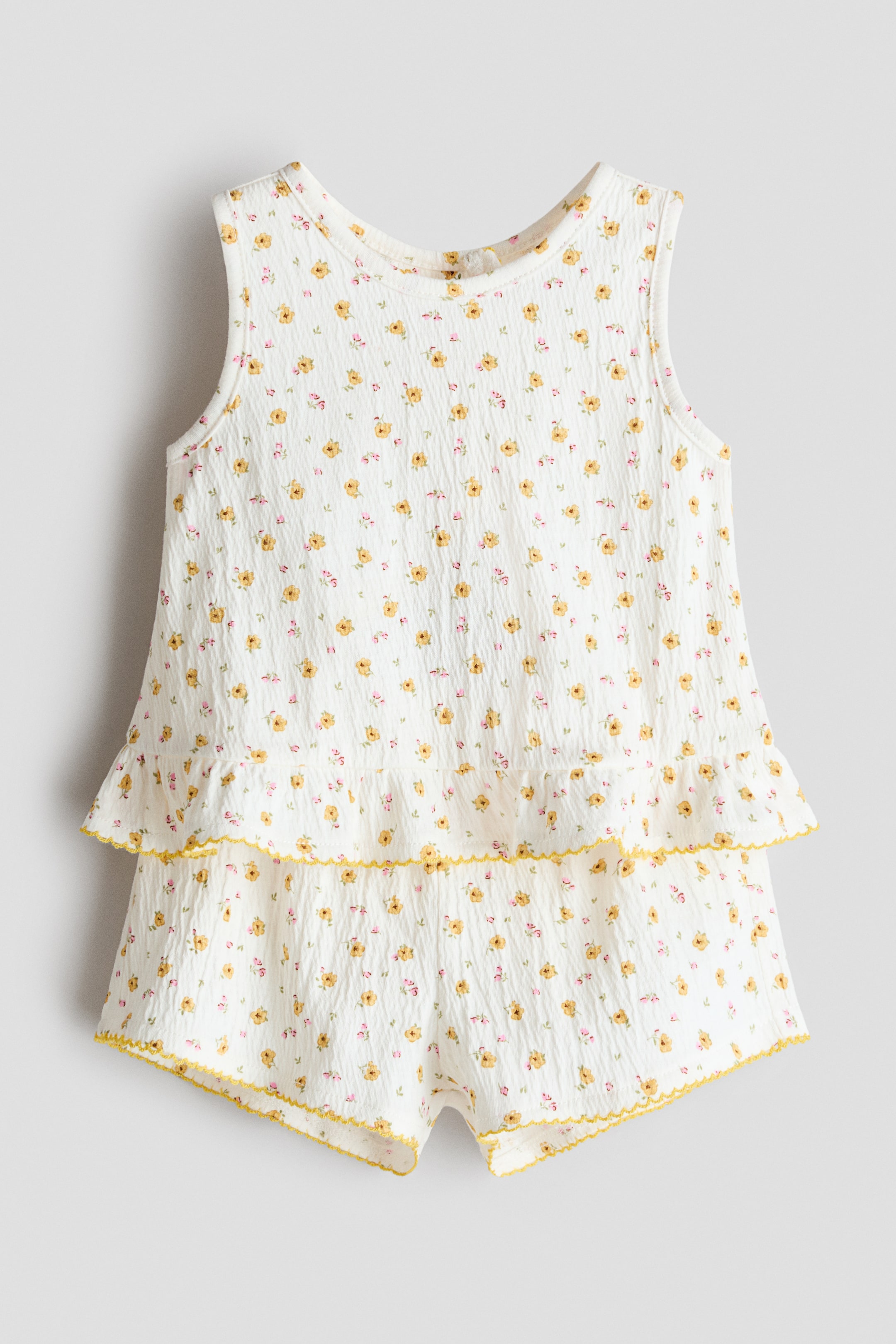 View larger image: This two-piece set, featuring a sleeveless top and shorts, is shown from the front, crafted from crinkled fabric with a small floral print. The top displays a round neckline and a gathered frill at the hem, while the shorts are visible below, both accented with yellow picot trim.