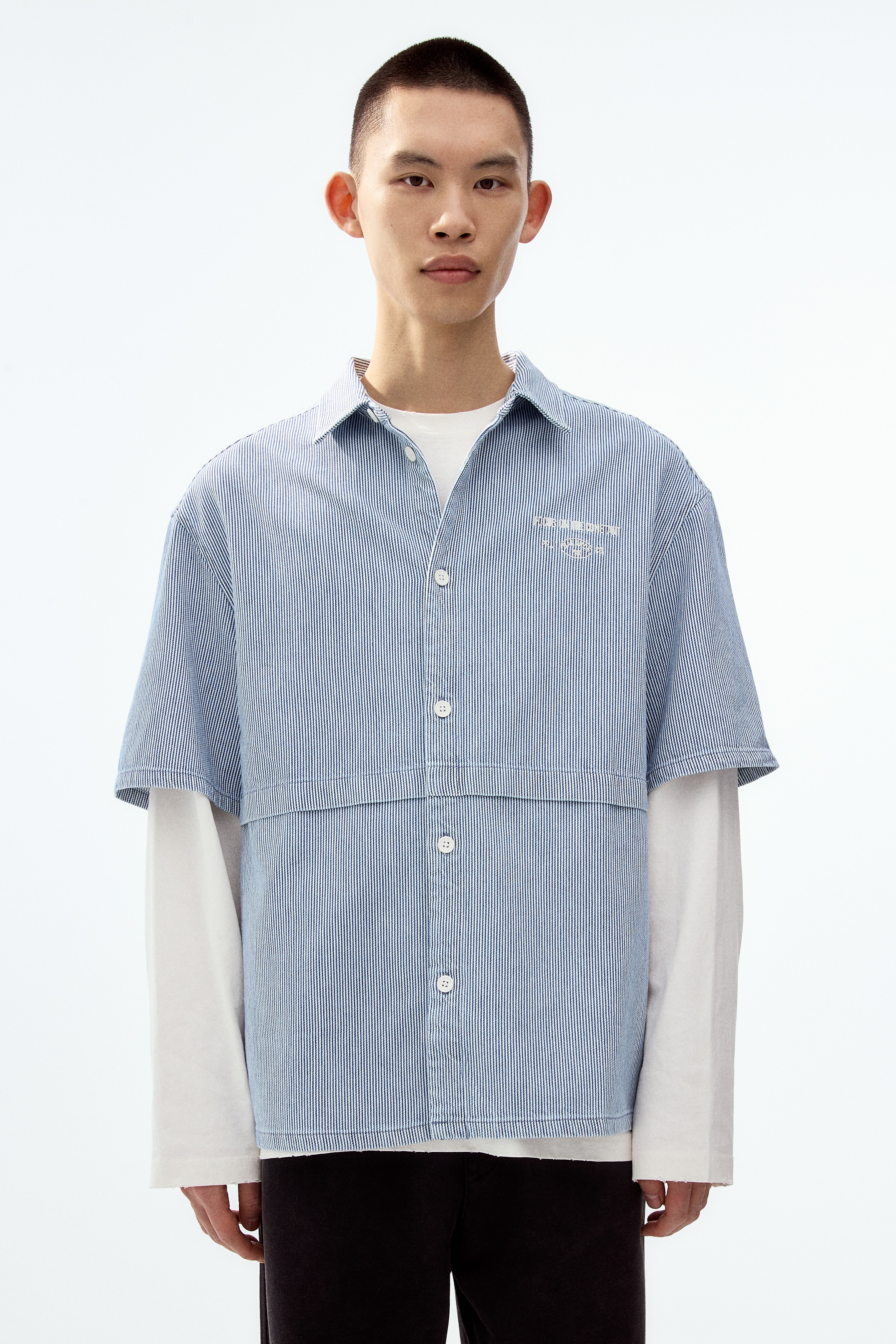 A light blue and white pinstriped short-sleeved shirt is presented from the front. It is displaying a pointed collar, a full-button front closure with white buttons, and white embroidered text on the left chest, while a horizontal seam is running across the chest, intersecting with a vertical seam that extends downwards from its center.