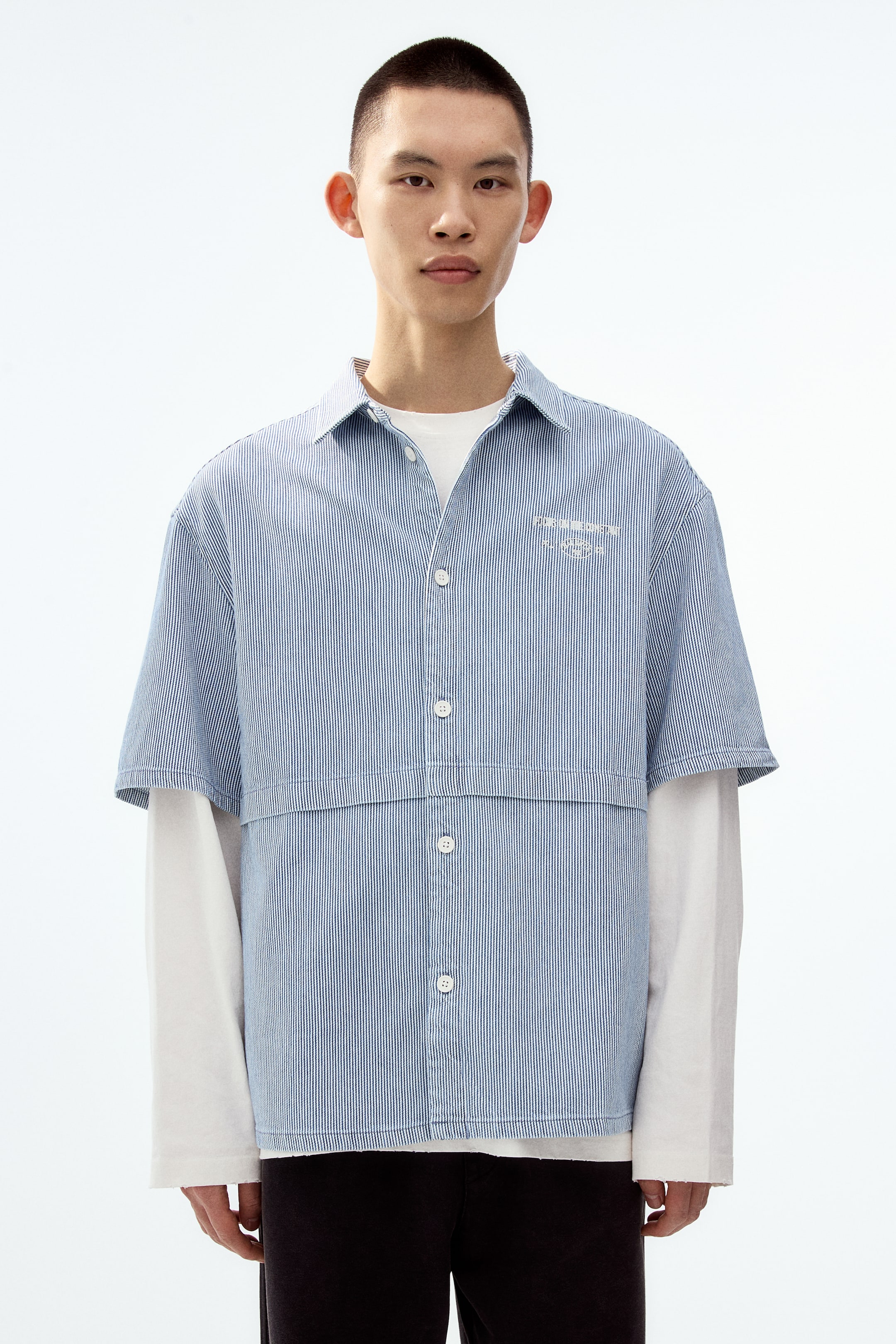 View larger image: A short-sleeved shirt in a blue-and-white pinstriped cotton weave is worn by the model. It features a turn-down collar, a classic button-up front, a horizontal front pleat, an embroidered motif on the chest, and a straight-cut hem that provides a loose fit. The model layers the shirt over a white long-sleeved shirt.