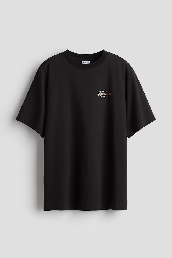 A black cotton-blend sports T-shirt is shown from the FRONT, featuring a relaxed fit with dropped shoulders and a straight-cut hem. It is designed with a round, rib-trimmed neckline and displays a golden script print reading "ALL/N Equestrian" on the front chest. - 1