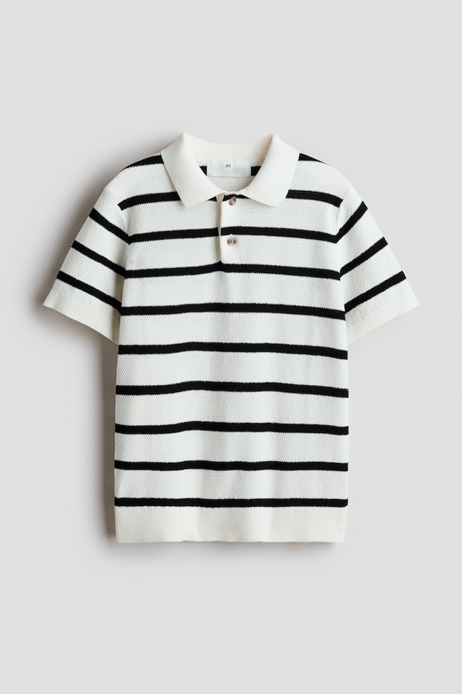 A white polo shirt with black horizontal stripes is shown from the front, featuring a waffle-knit texture, a collar, a two-button placket, dropped shoulders, short sleeves with ribbed cuffs, and a ribbed hem. - 1