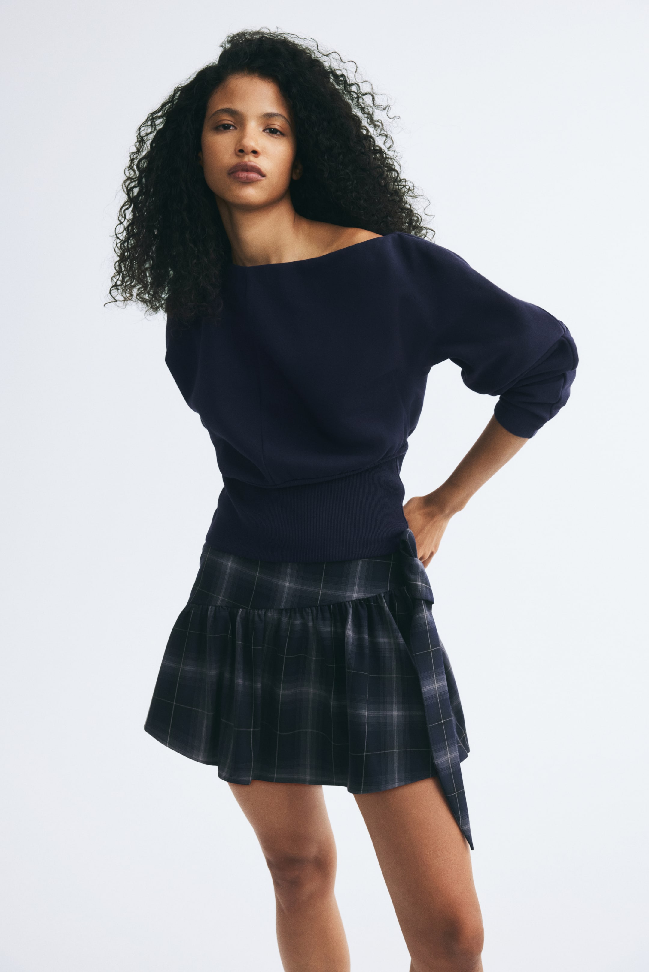 Größeres Bild ansehen: Ein marineblauer Sweatshirt mit U-Boot-Ausschnitt und breitem Rippbund wird vom Model getragen und mit einem dunklen karierten Minirock kombiniert, der mit einem gerafften Saum und seitlicher Bindung versehen ist. Das Model posiert mit einer Hand auf der Hüfte und setzt so den Fall und die Passform der Kleidungsstücke in Szene.