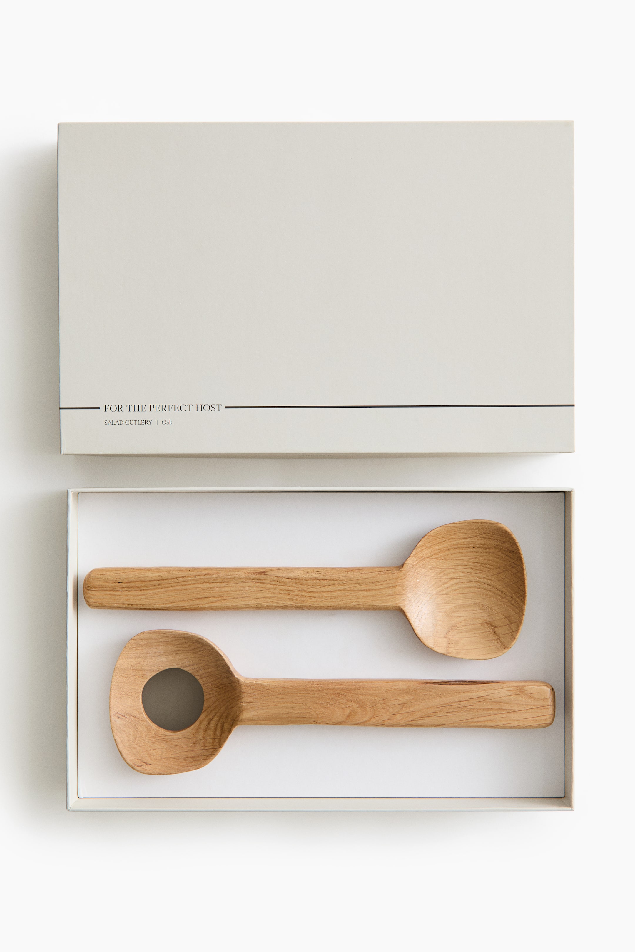 View larger image: A wooden salad cutlery set is resting inside an open, light beige gift box, with the lid positioned above. The set features one solid wooden serving spoon and one serving spoon with a circular cut-out, both showing natural oak wood grain and flat, straight handles.