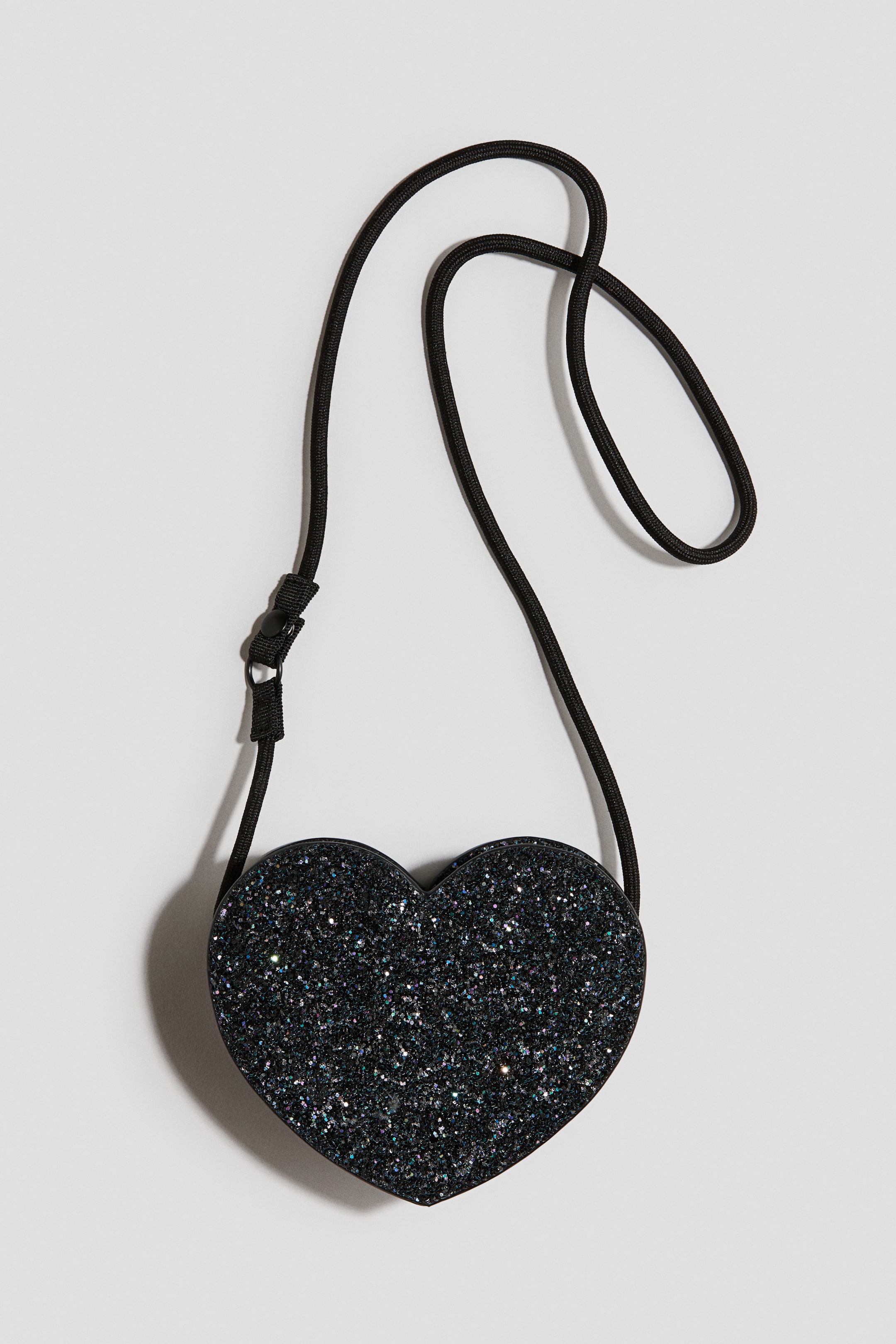 Ver imagen más grande: Un bolso bandolera negro en forma de corazón, con brillo, se muestra de frente. Presenta un cierre de cremallera en la parte superior y una correa de hombro fina de cordón negro con un cierre a presión separable en un extremo.
