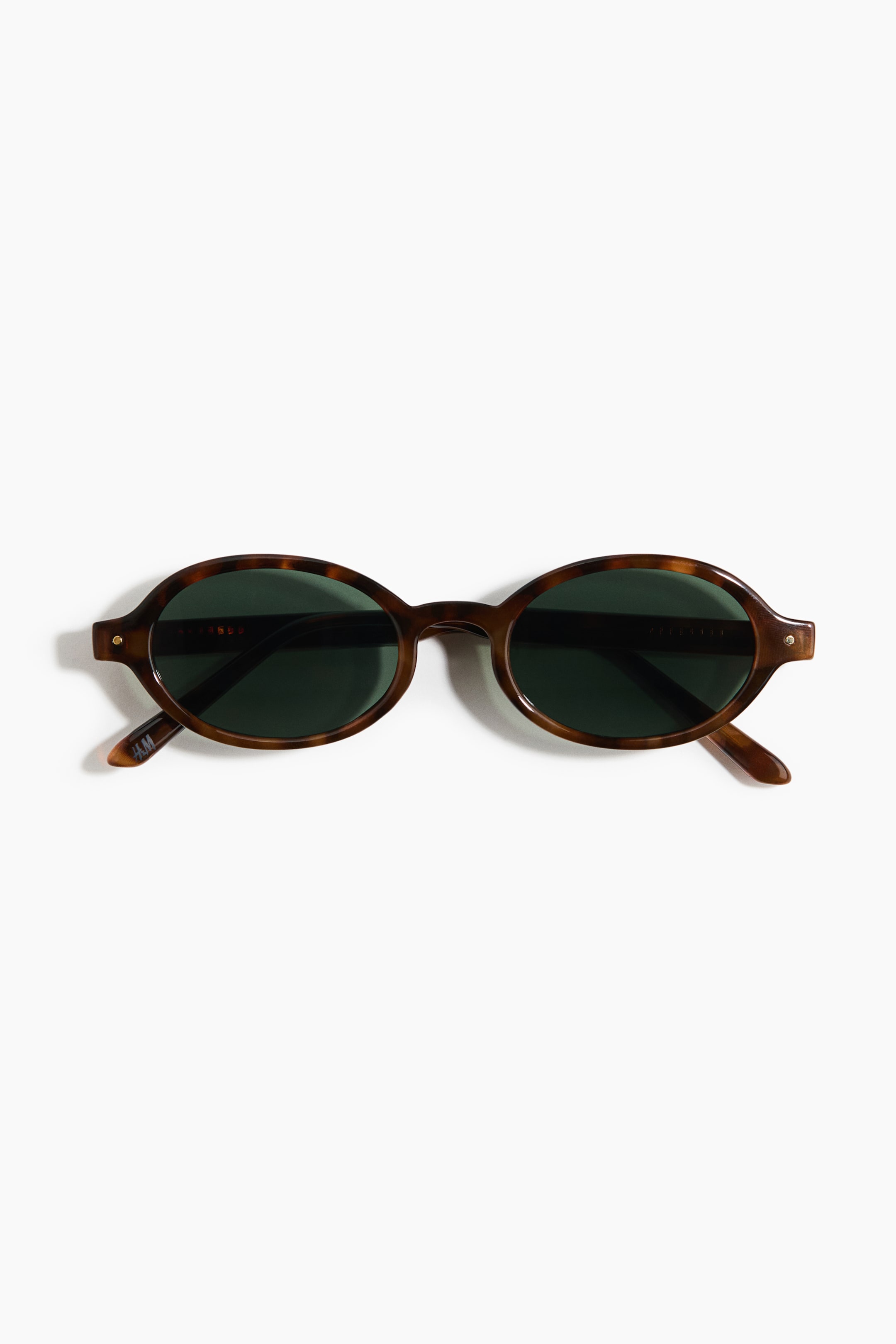 View larger image: The sunglasses are positioned front-facing, showcasing oval tortoiseshell frames paired with dark green lenses. Small metallic pins accentuate the front corners of the frame.
