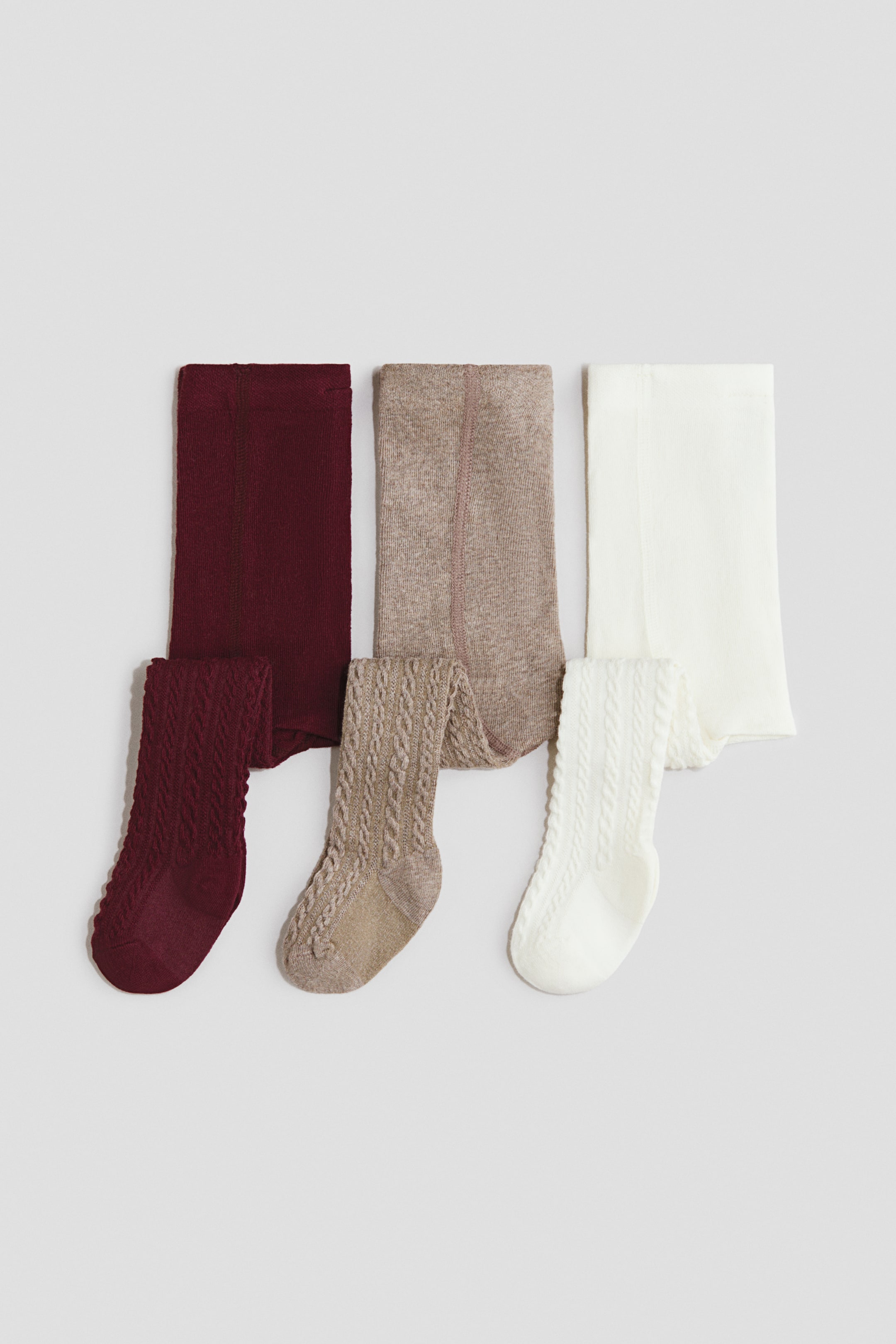 View larger image: Three pairs of tights are laid flat, partially folded. One pair is deep red, one is beige, and one is off-white, each featuring a smooth knit on the upper section and a cable knit pattern on the leg and foot.