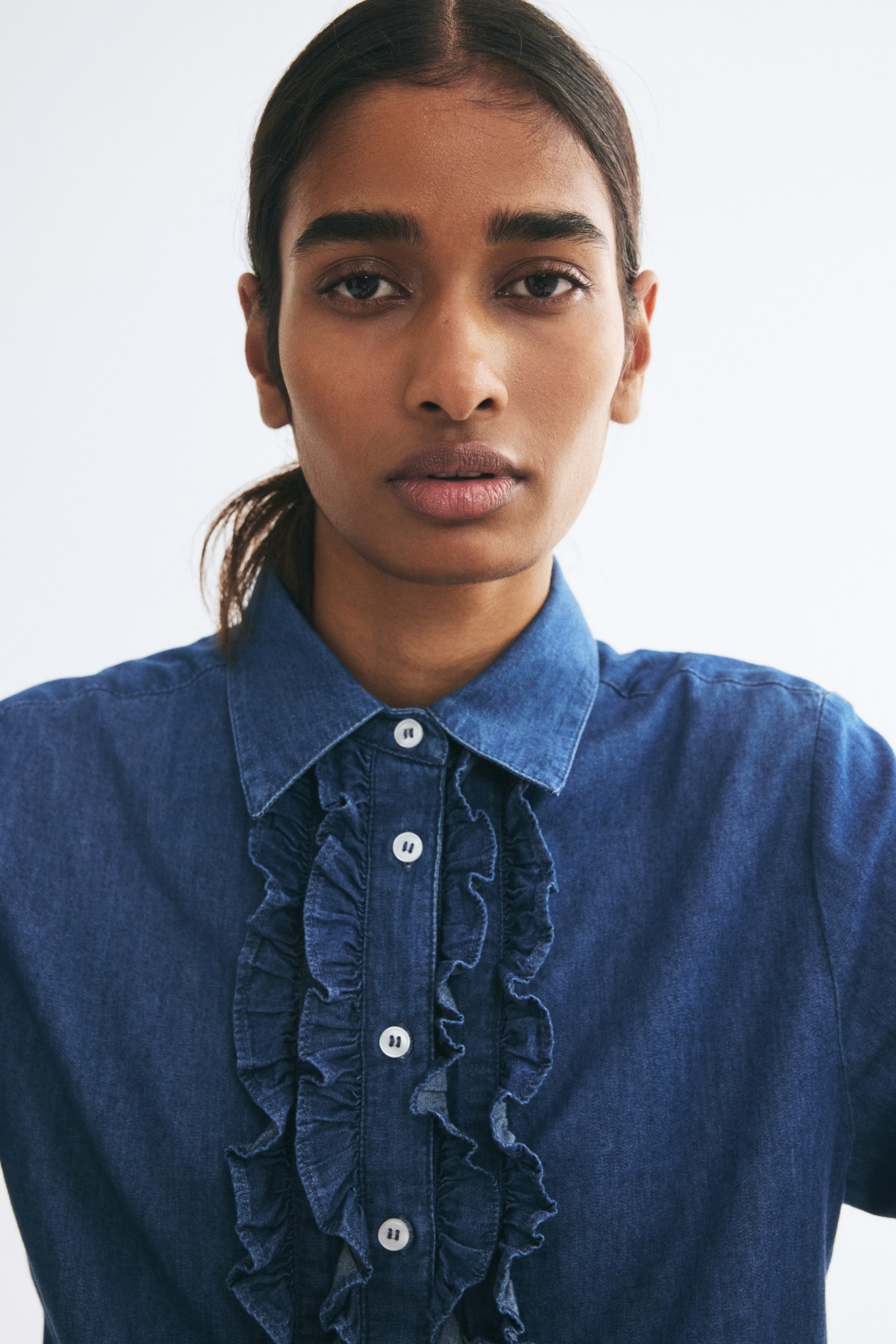 View larger image: The model is wearing a blue denim shirt featuring a pointed collar and a button-down front with a frill-trimmed placket. The close-up shot highlights the white buttons and ruffled detailing along the centre band.