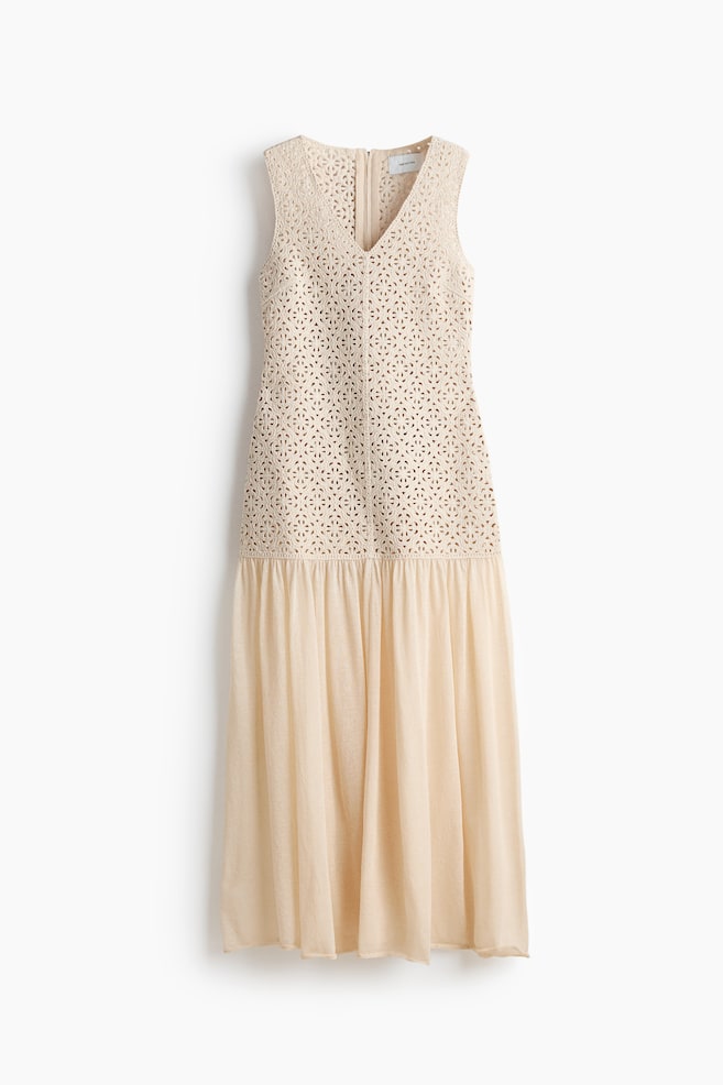 A light beige sleeveless maxi dress is displaying its back. The bodice is featuring an intricate cutwork pattern with a V-back and a visible zipper closure. The skirt is gathering at the waist, flowing into a long, soft drape. - 1