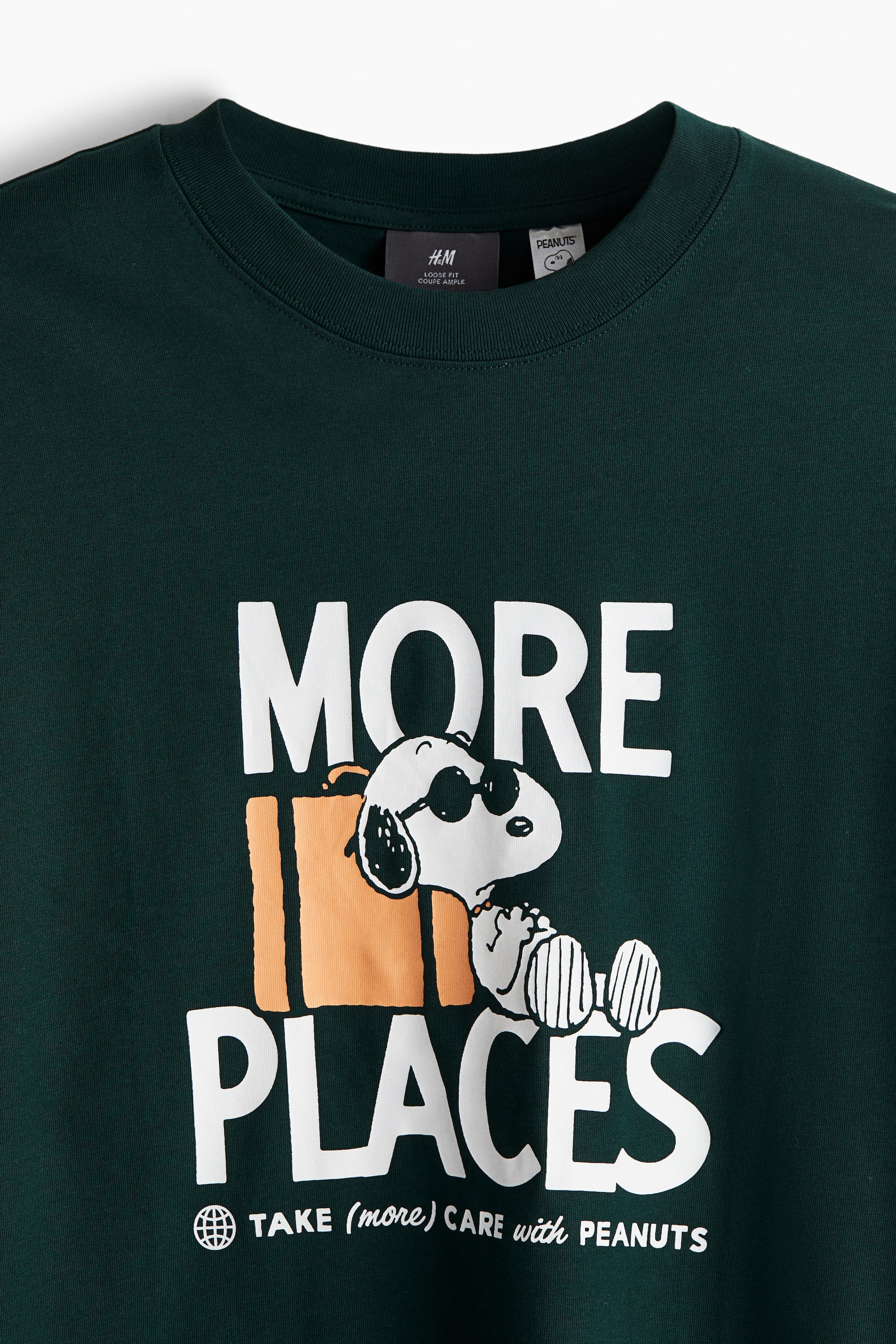 View larger image: A dark green T-shirt is displaying a large graphic print on its FRONT, which is featuring Snoopy wearing sunglasses and leaning against an orange suitcase, positioned between the white words "MORE" and "PLACES." The T-shirt is designed with a round neckline and a narrow ribbed trim, and product tags are visible at the back of the neck.