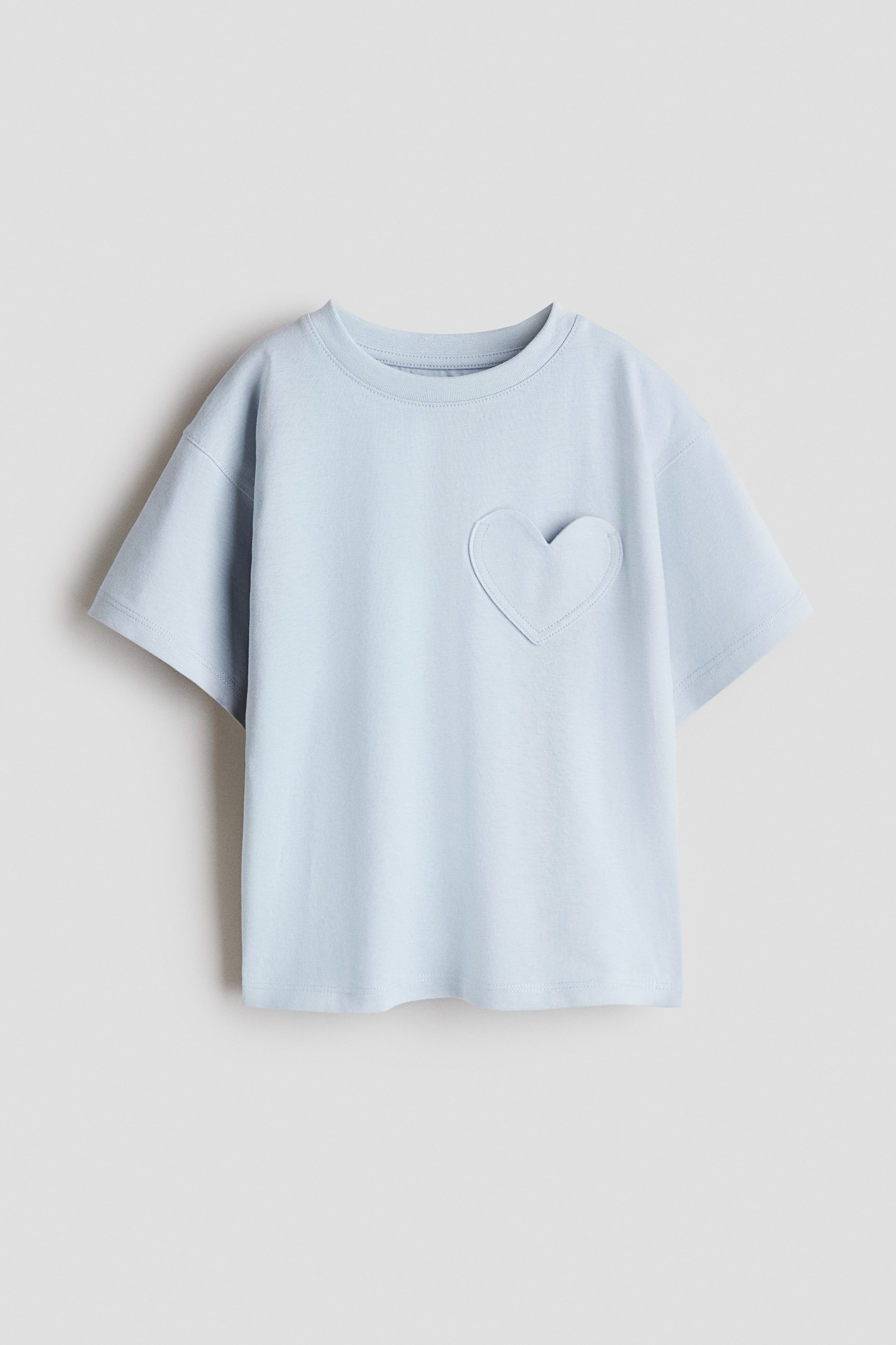 View larger image: A light blue t-shirt is lying flat, featuring a ribbed crew neck and short sleeves. A heart-shaped appliqué, outlined with visible stitching, is attached to the left chest.