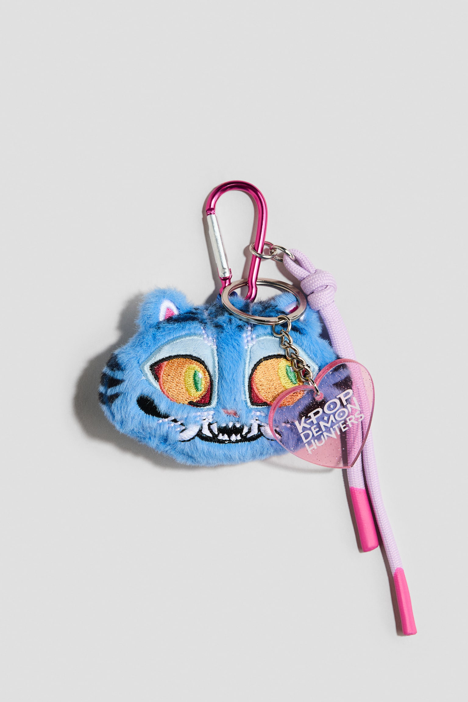 A plush bag charm features a blue furry tiger head with large, colourful embroidered eyes and a toothy grin. It hangs from a pink carabiner hook, accompanied by a translucent pink heart-shaped charm displaying "K-POP DEMON HUNTERS" and a knotted lavender cord with pink-dipped ends. - 1