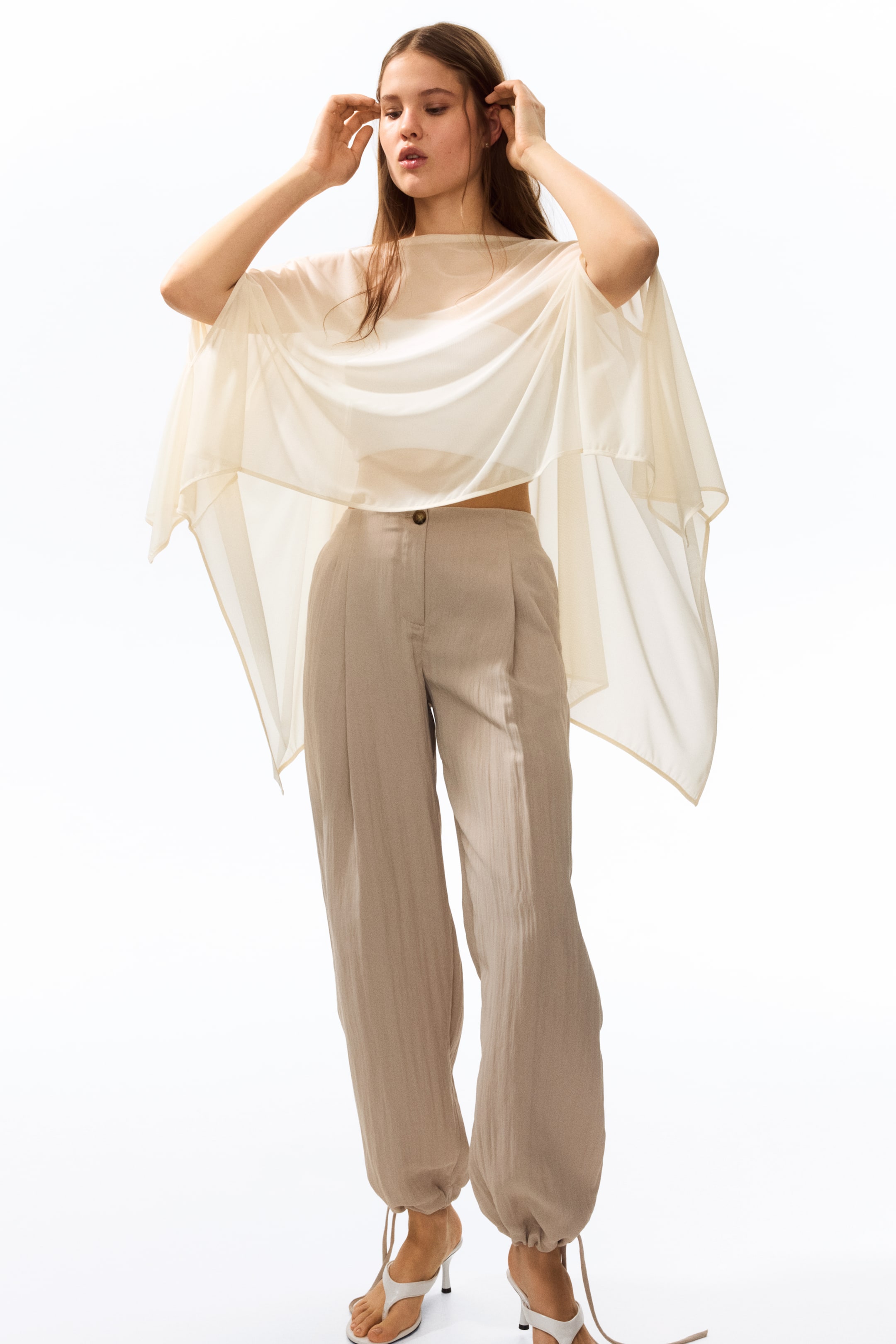 Ver imagem maior: Uma capa de malha transparente de cor creme com decote barco está a ser usada pela modelo sobre uma blusa leve. O seu tecido em camadas cai suavemente em torno dos ombros e do corpo, terminando acima da linha da cintura.