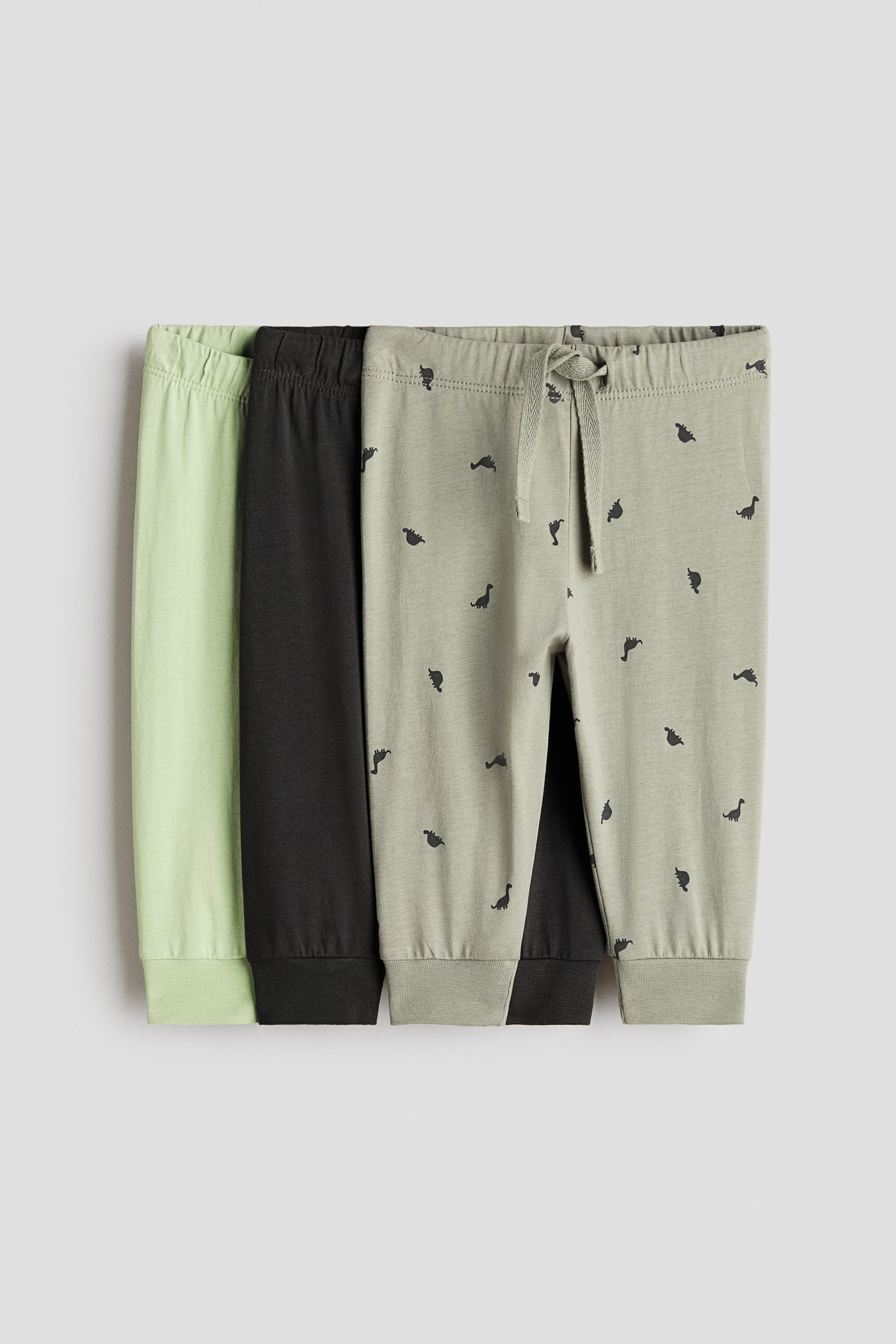 View larger image: Three pairs of joggers are lying flat, presenting their front sides, each featuring an elasticated waistband and straight legs ending in ribbed cuffs. The frontmost sage green jogger is displaying a drawstring tie and a repeating black dinosaur print, while the other two are plain black and light green.
