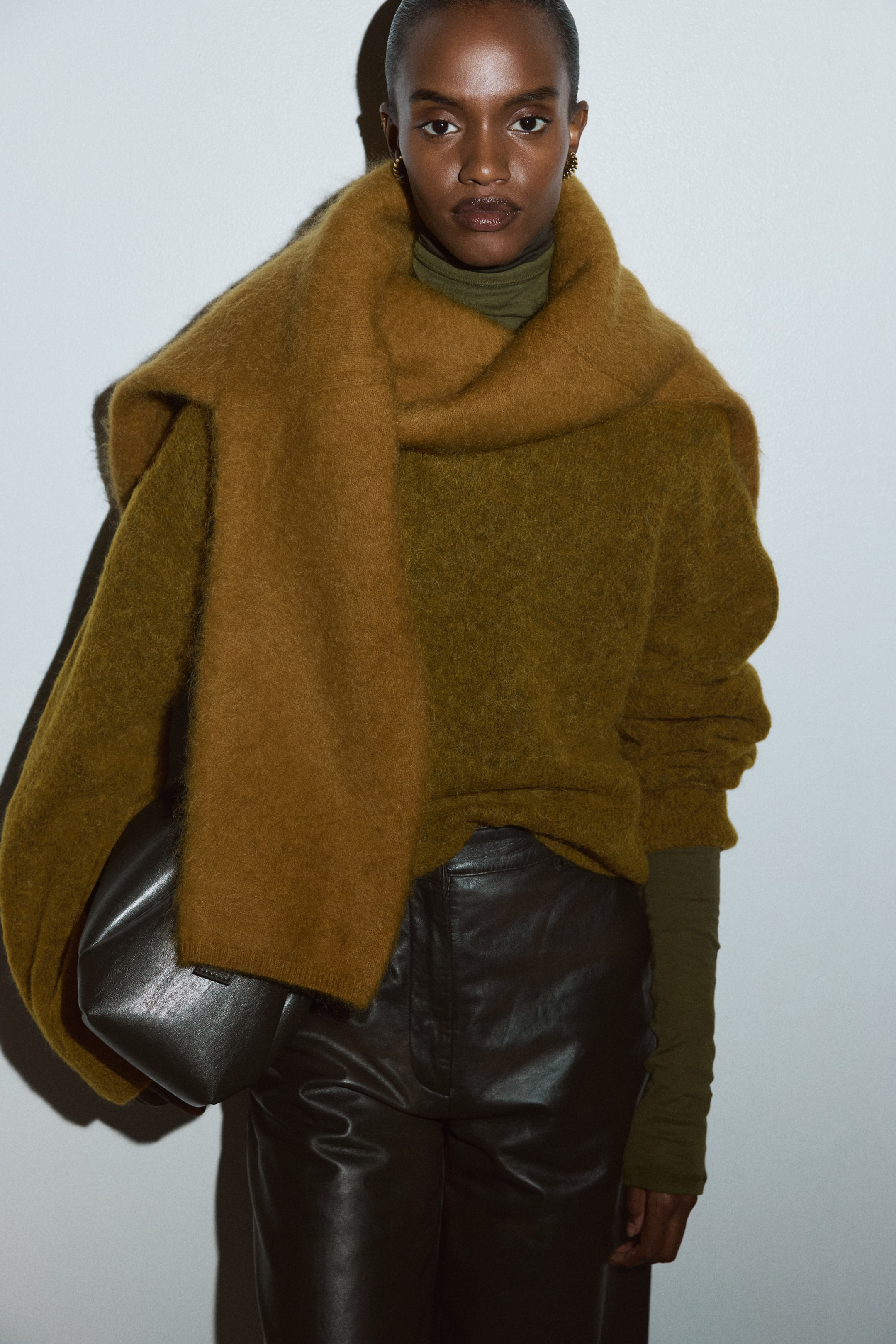 Agrandir l'image: Une écharpe chaude et marron, duveteuse est drapée sur un pull duveteux vert olive, qui est porté sur un col roulé vert olive plus clair. Un pantalon en cuir marron foncé est visible en dessous, avec un sac doux gris foncé tenu contre la hanche.