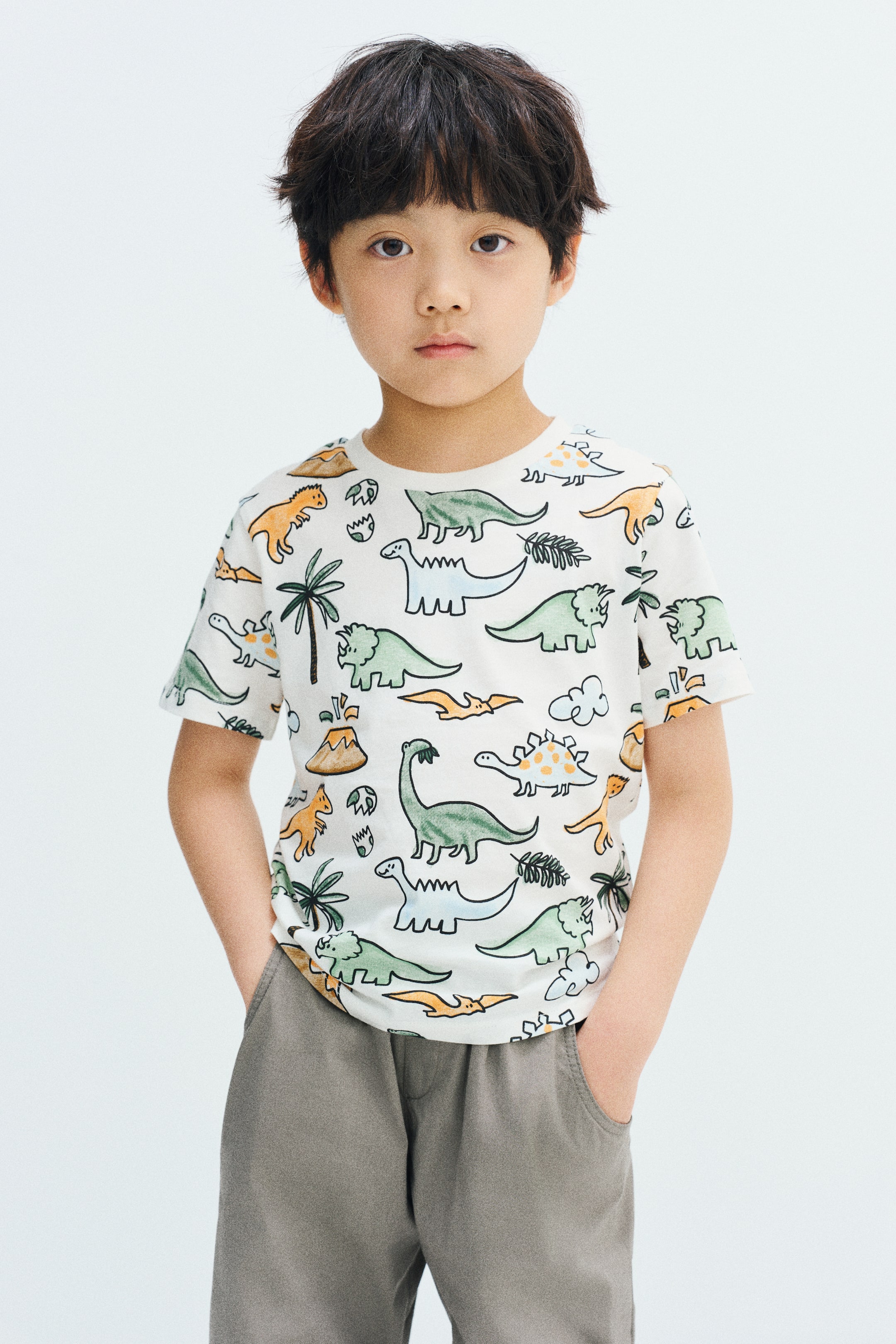 View larger image: The model is wearing a white, short-sleeved cotton jersey T-shirt featuring an all-over colorful dinosaur print and a round, rib-trimmed neckline. The model is posing with hands placed in the pockets of grey trousers.