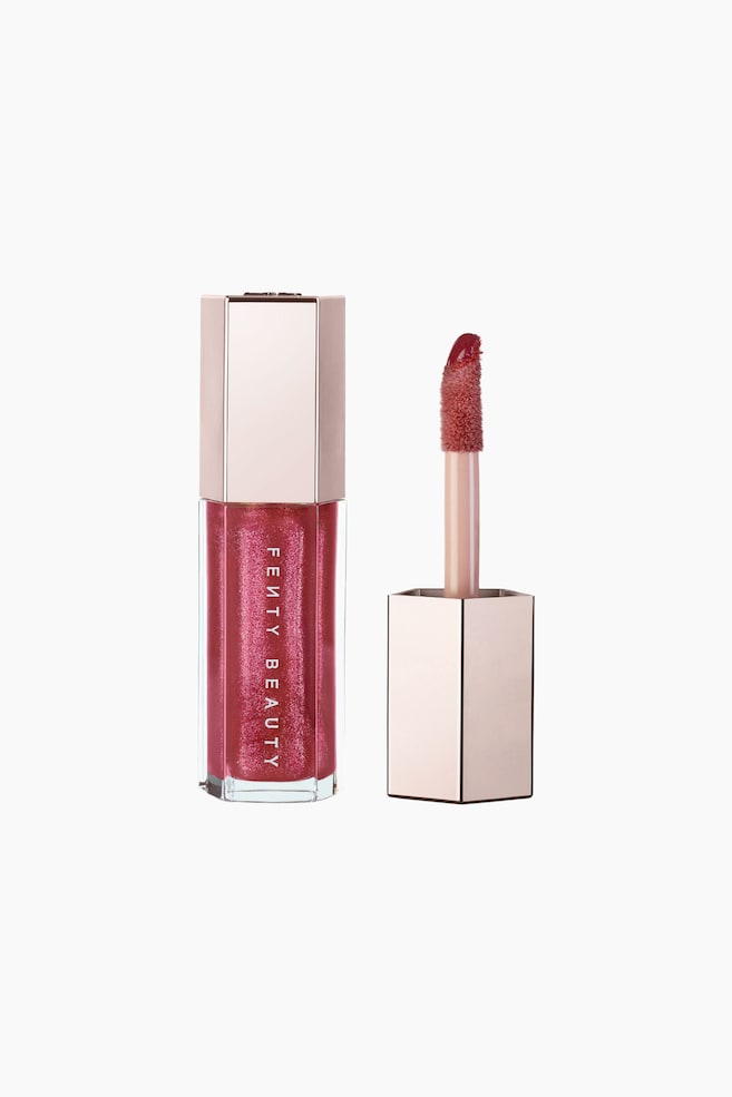 En hexagonal Fenty Beauty-lipglossflaske i en skinnende rosapink nuance stÃ¥r oprejst ved siden af sin afskruede applikatorstav. Den gennemsigtige flaske viser lodret hvid branding, mens staven har en stor doe-foot-applikator dÃ¦kket af den blanke formel under en metallisk rosaguld hÃ¦tte. - 1