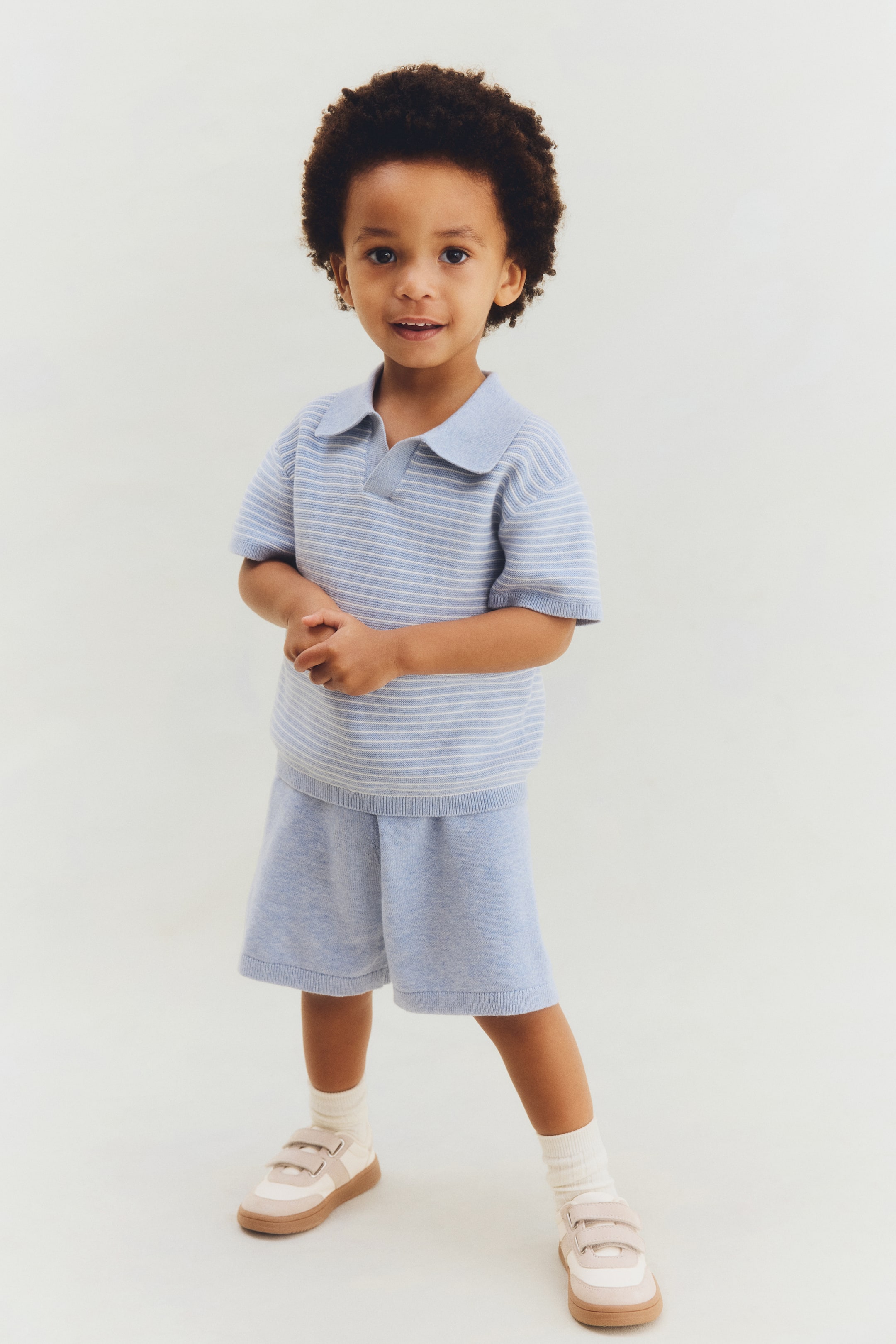 Grotere afbeelding bekijken: Een gebreide tweedelige outfit bestaande uit een lichtblauw poloshirt met korte mouwen en witte horizontale strepen, gecombineerd met bijpassende lichtblauwe shorts, getoond vanaf de voorkant.