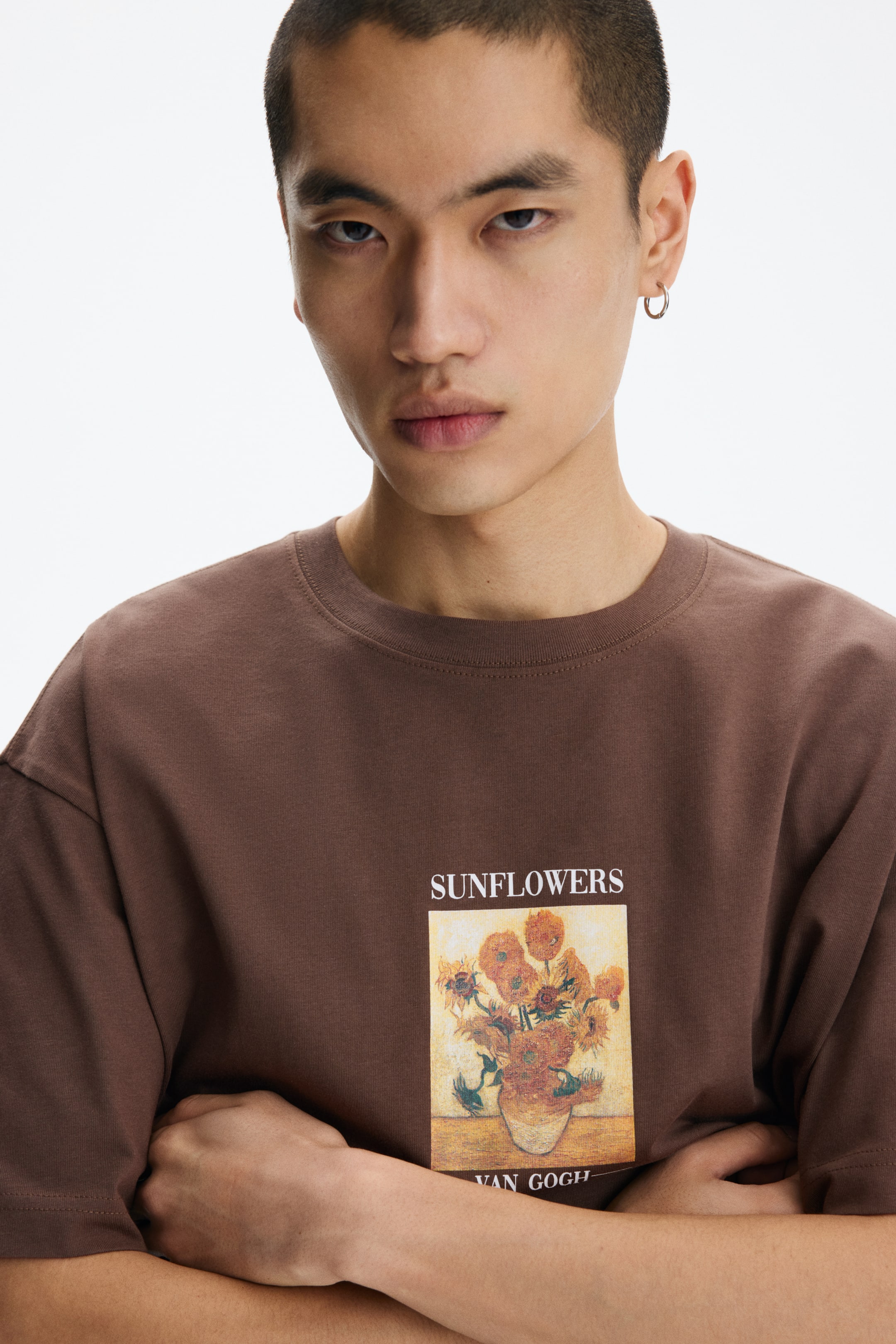 Agrandir l'image: Un t-shirt à col rond marron foncé présente des manches courtes et un col côtelé. Sur le devant, un imprimé graphique rectangulaire représente le tableau "Tournesols" de Van Gogh, avec "TOURNESOLS" écrit au-dessus et "VAN GOGH" en dessous.