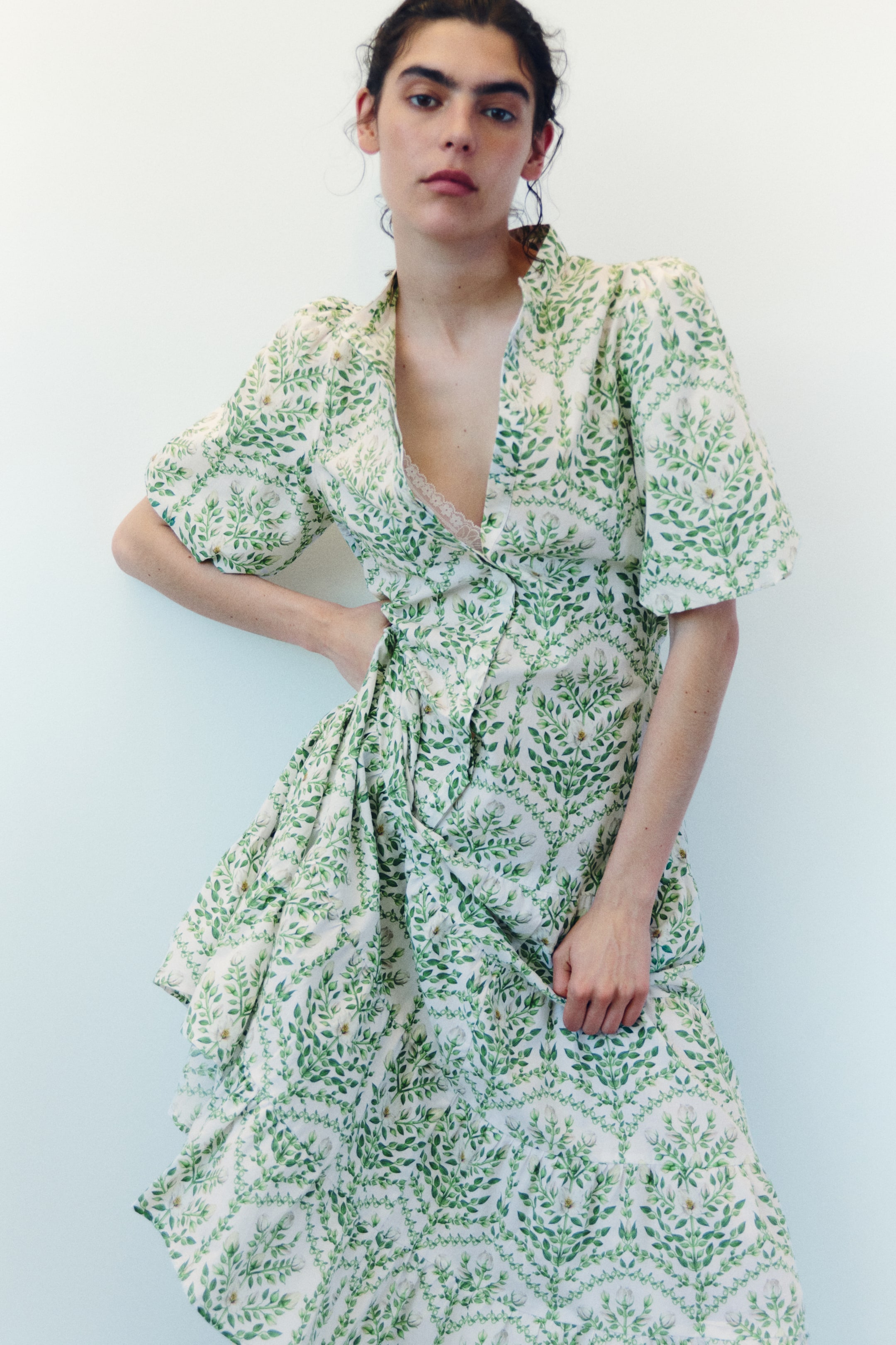 Visa större bild: En vit omlottklänning visar ett upprepat mönster av gröna blad och små vita blommor, med en djup V-ringning, korta puffärmar och en draperad, rynkad kjol. En vit spetsbralette syns under V-ringningen.