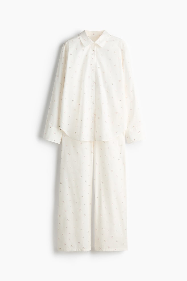 Pyjama set shown FRONT, consisting of a cream-coloured shirt and matching bottoms in a soft cotton weave with a small floral pattern. The shirt features a collar, buttons down the front, dropped shoulders and long sleeves with a slit and button at the cuffs, together with rounded side vents at the hem, while the bottoms have an elasticated, drawstring waist. - 1