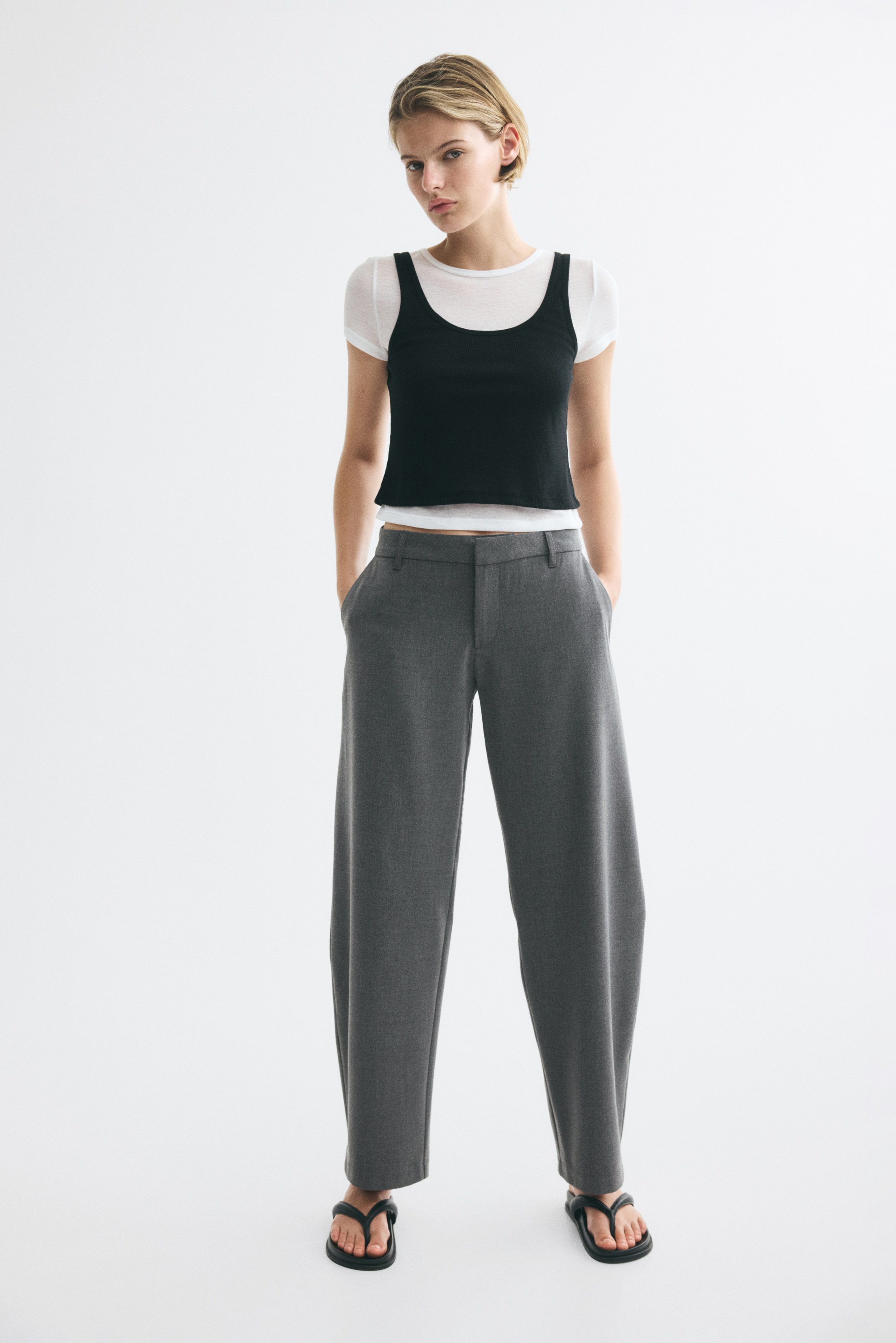 Dark grey trousers are displayed from the front, featuring a flat waistband with belt loops and slanted side pockets. The trousers show straight legs and a plain hem.