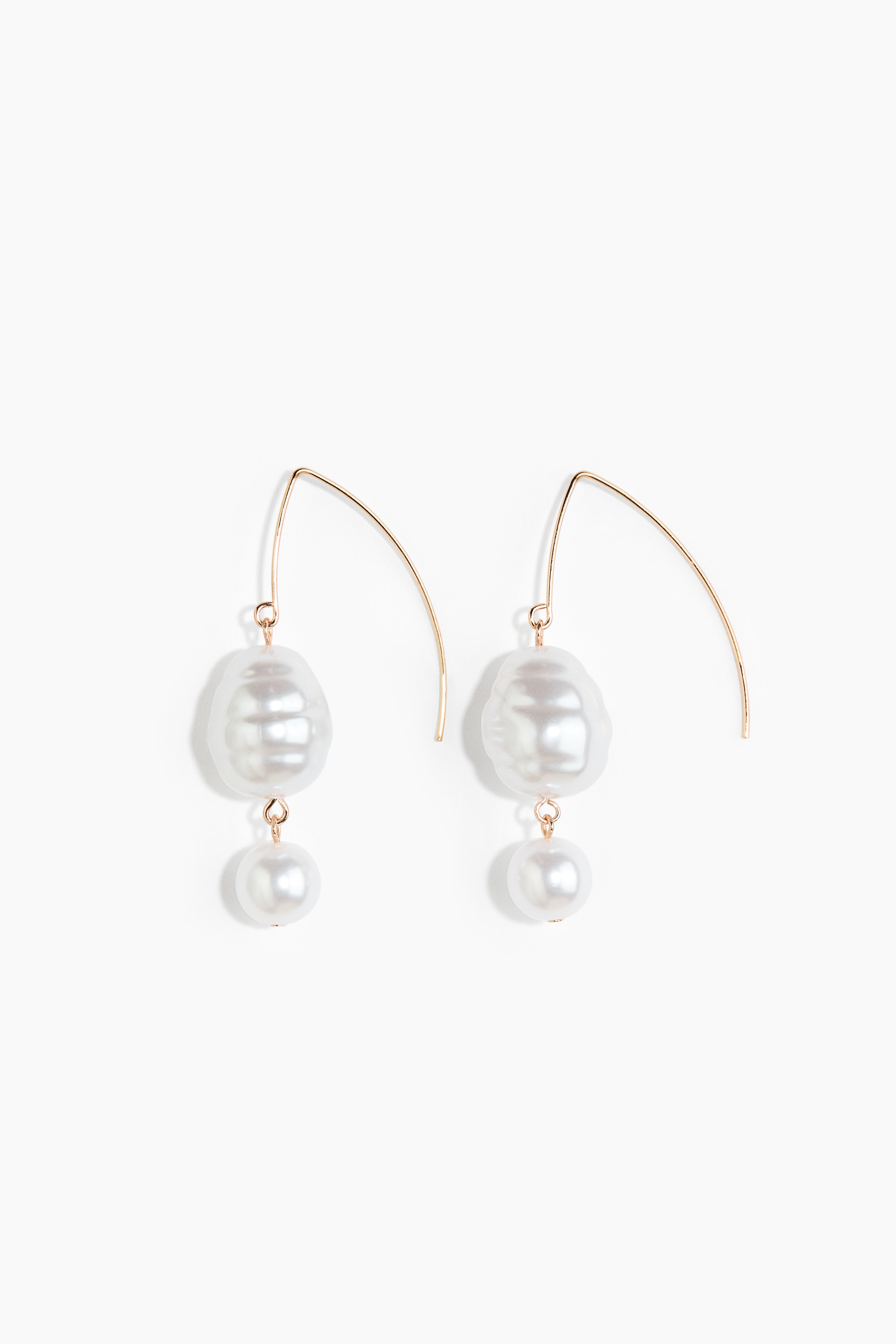 The model is wearing metal pendant earrings featuring pearly plastic beads in different sizes. The visible earring features a curved gold-tone hook suspending a large, irregular baroque-style bead and a smaller round bead. The jewellery is styled with a high-collared, cream-coloured garment featuring lace-up back details.