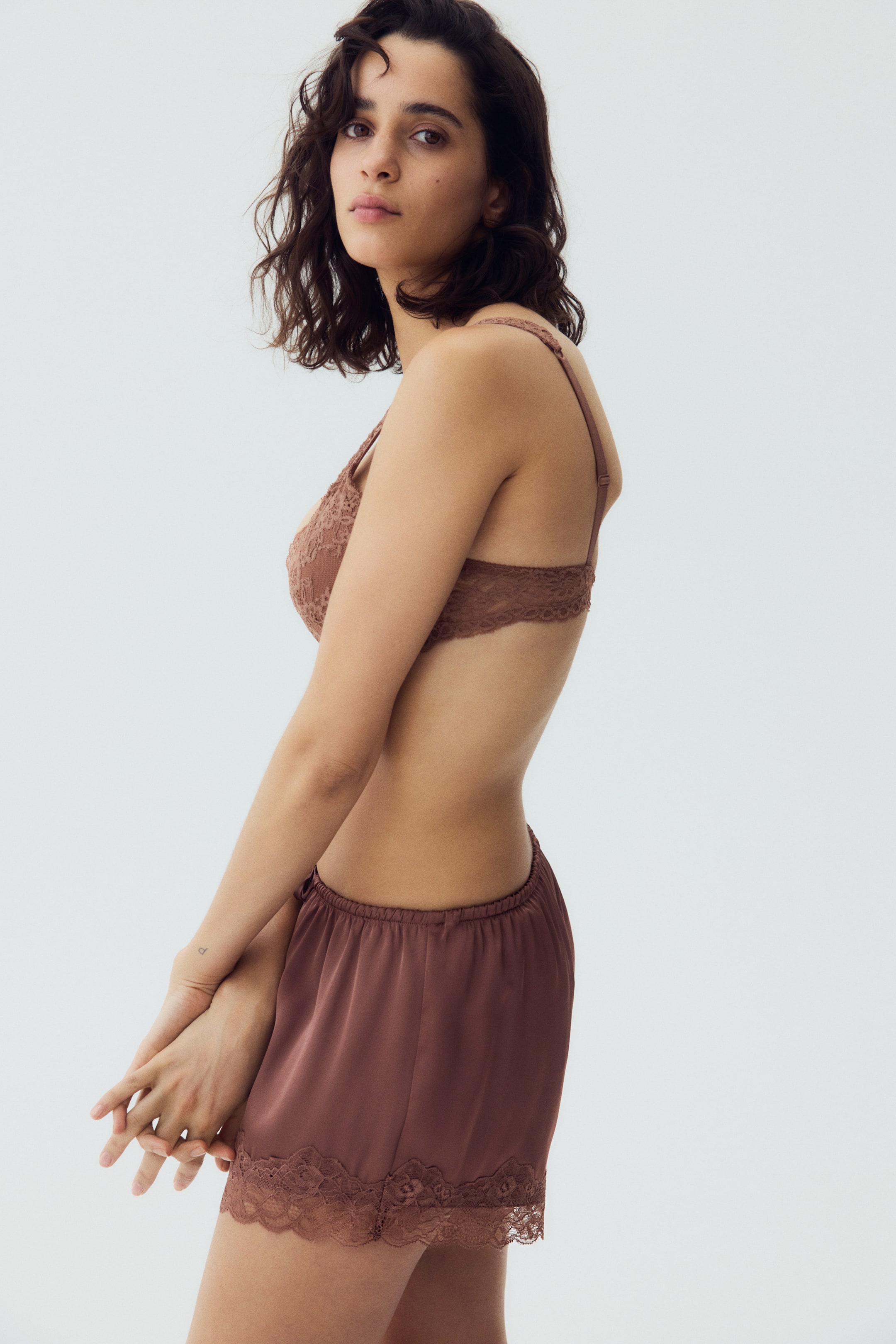 Ver imagem maior: Um bralette de renda rosa empoeirada apresenta alças ajustáveis e uma faixa de renda larga, coordenando com calções de seda a condizer. Os calções apresentam uma cintura elástica e um acabamento de renda intrincado ao longo da bainha, com todo o conjunto voltado para um ângulo lateral.