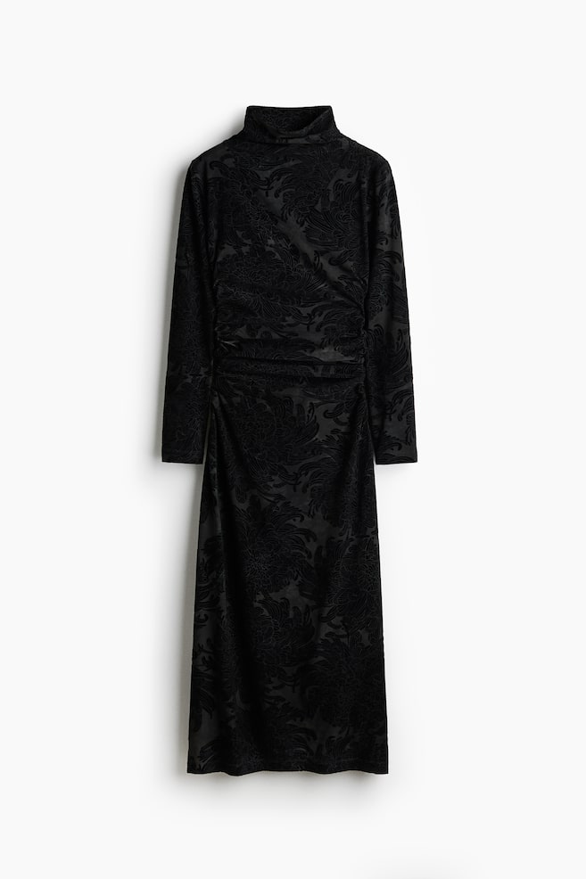 Un vestido negro de manga larga está colgado, mostrando su vista frontal, con un cuello alto y un patrón floral y ornamental en relieve a lo largo de su tela similar al terciopelo. Se observa fruncido a lo largo del lado izquierdo, creando pliegues desde la cintura hasta el largo midi, en una silueta de ajuste ceñido. - 1