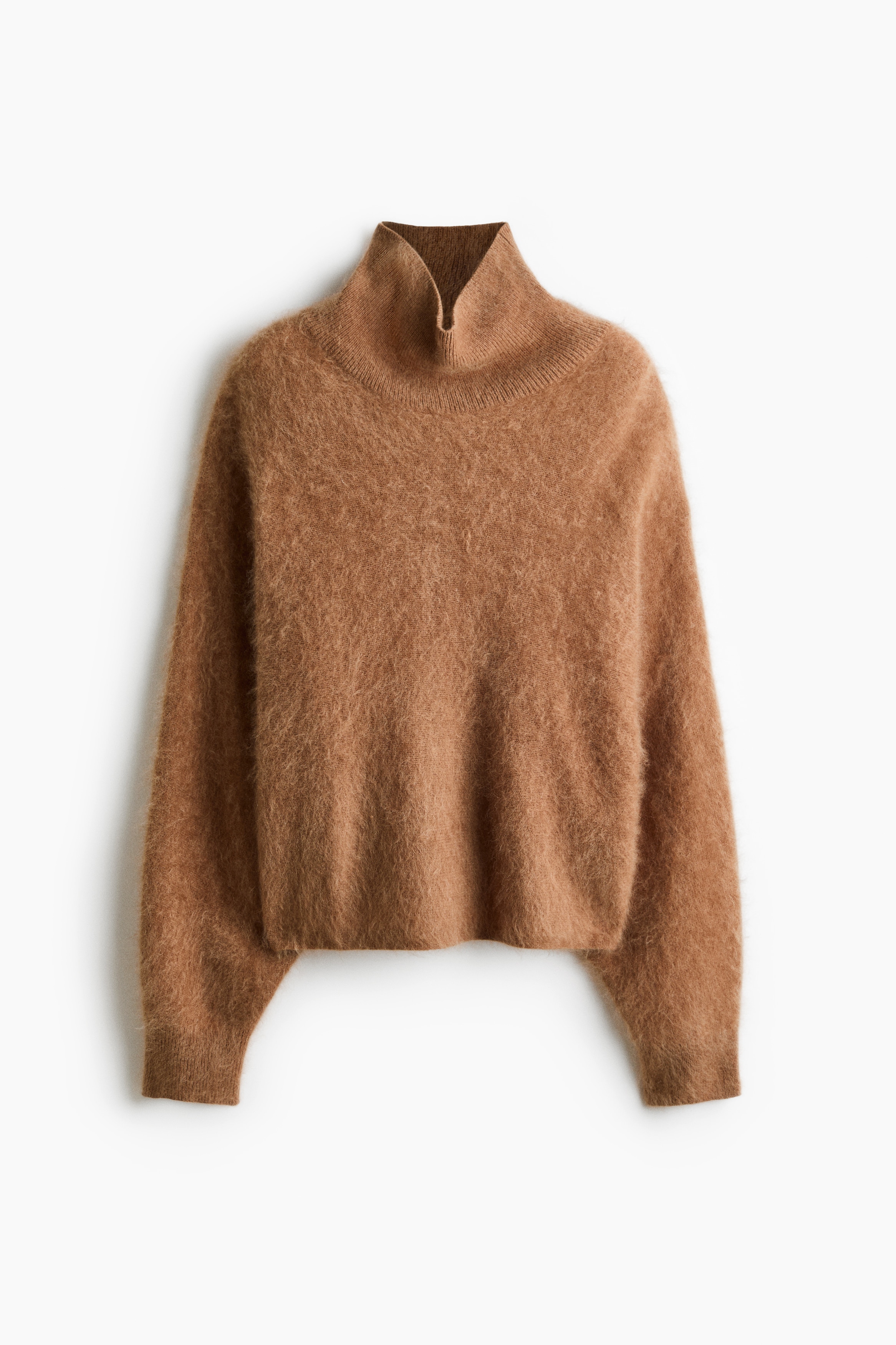 The model is wearing an oversized brown sweater in a brushed wool knit, featuring a high ribbed neck and long, voluminous sleeves. A matching knit garment is draped over the model’s shoulders, and the sweater is styled tucked into wide-leg white trousers.
