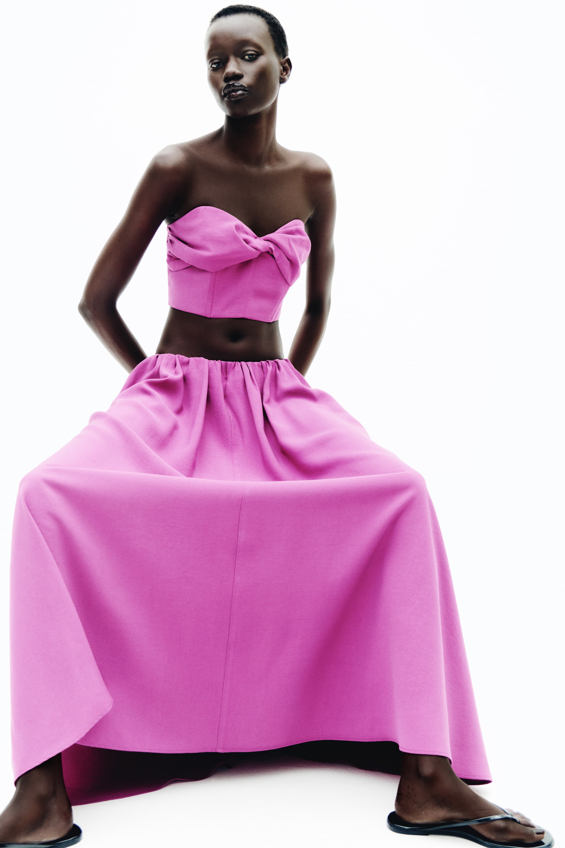 Grösseres Bild ansehen: Ein pinkes Bandeau-Crop-Top mit Sweetheart-Ausschnitt, das in der Mitte des Brustbereichs mit einer gedrehten Detailverarbeitung und plissierten Seiten versehen ist, wird vom Model getragen. Das Oberteil ist mit einem passenden pinken, weit ausgestellten Rock kombiniert.