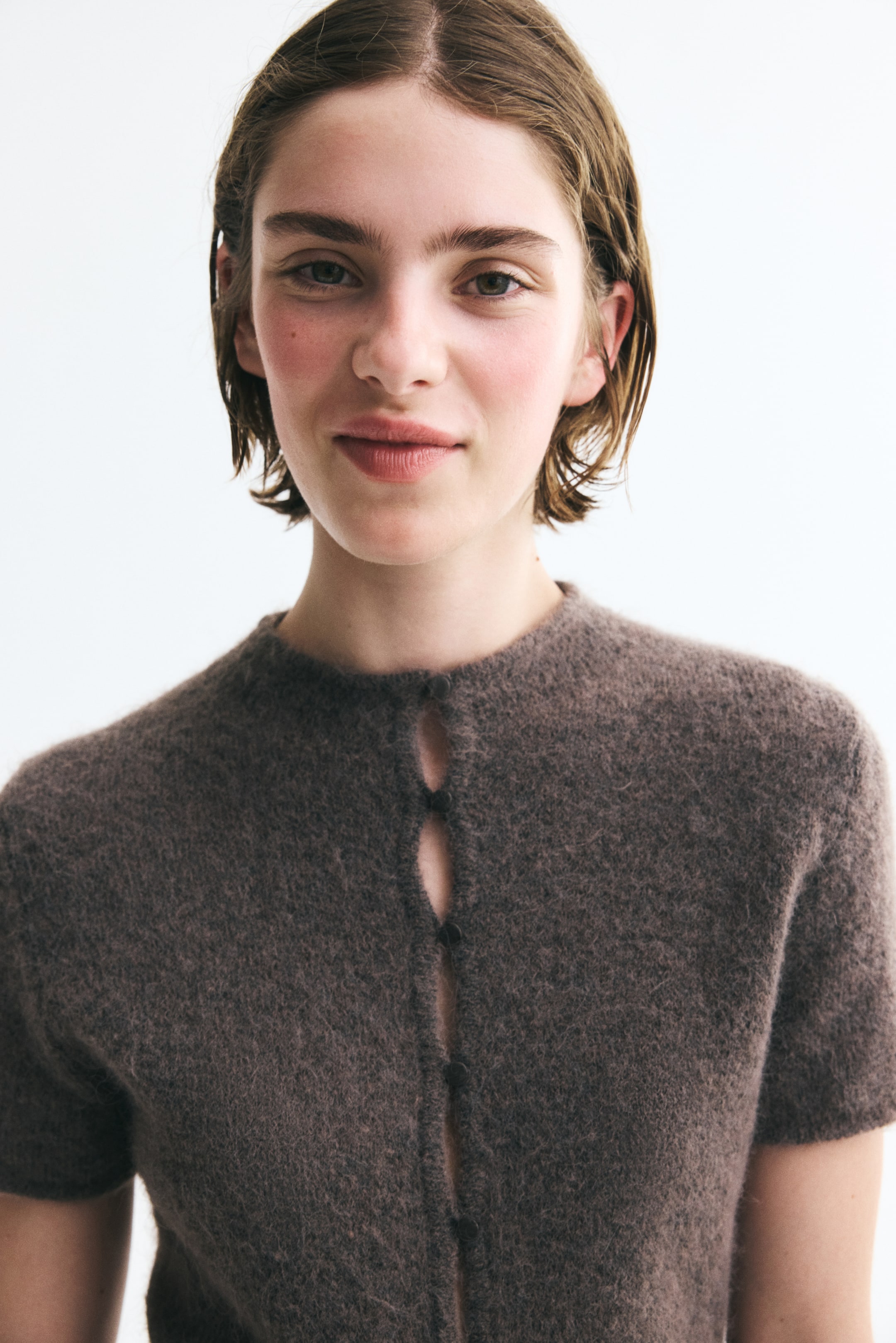 View larger image: A brown, short-sleeve cardigan in a fuzzy, fine knit is being worn by the model. The garment features a round neckline and small button-and-loop fasteners down the front.