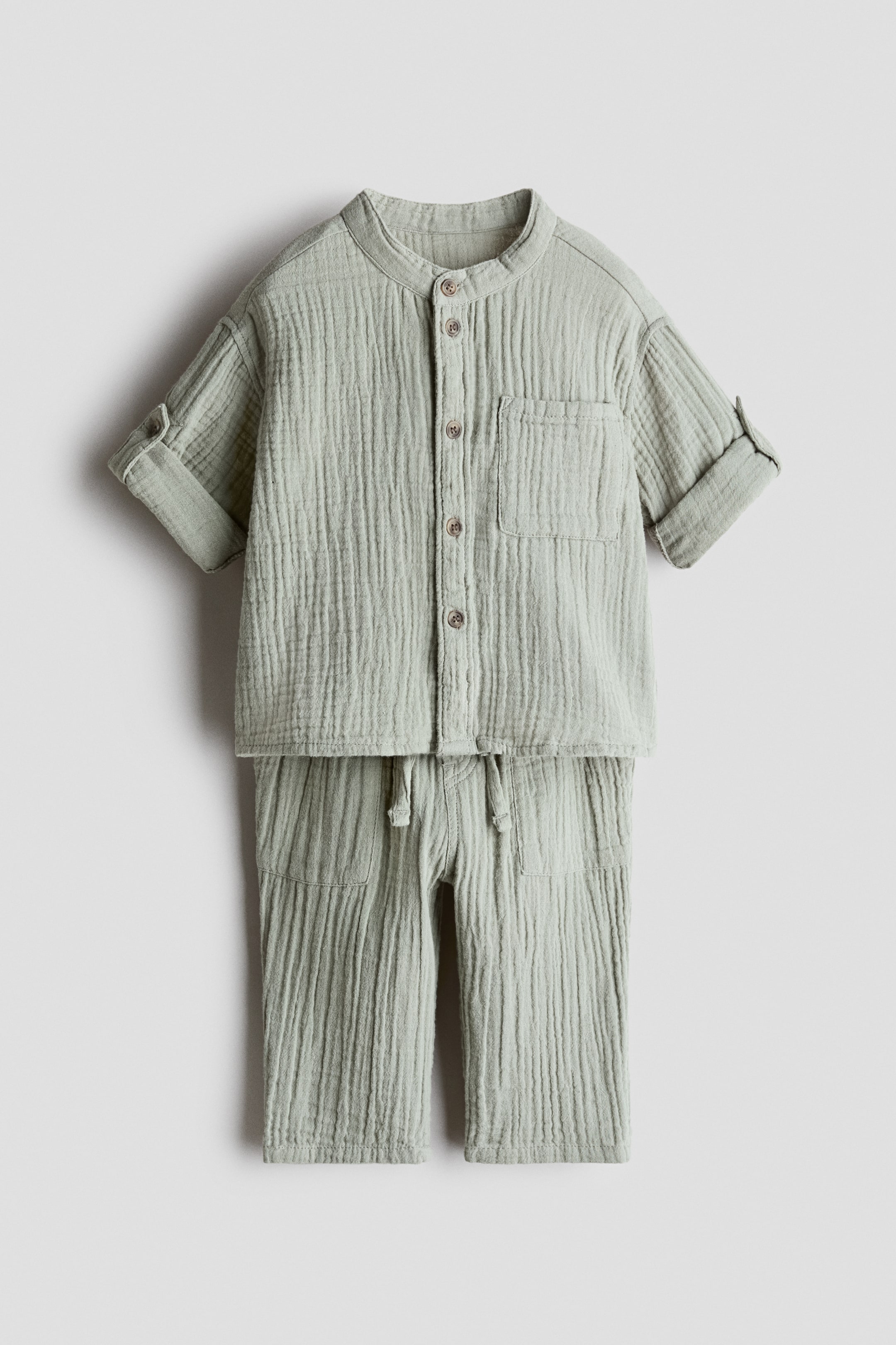 Voir une image plus grande: L'ensemble en mousseline de coton froissé vert clair, vu de FACE, comprend une chemise avec un col mao, des boutons sur le devant, une poche poitrine ouverte et des manches longues retroussées et maintenues par une patte et un bouton. Le pantalon assorti présente une taille élastiquée avec cordon de serrage noué et des poches latérales.