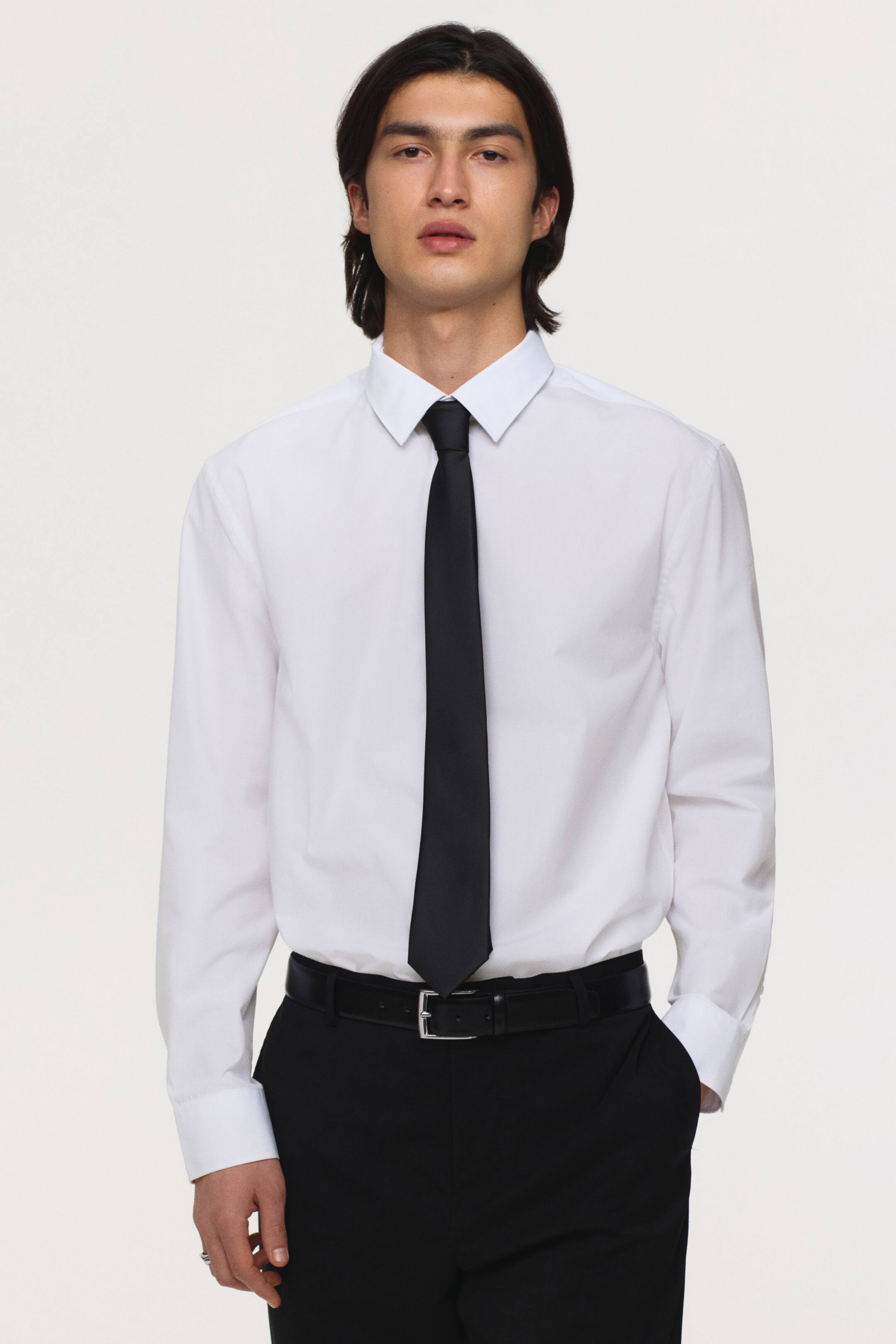 View larger image: A white dress shirt showcases a classic collar and long sleeves with buttoned cuffs. It is paired with a black tie, black trousers, and a black belt with a silver buckle.