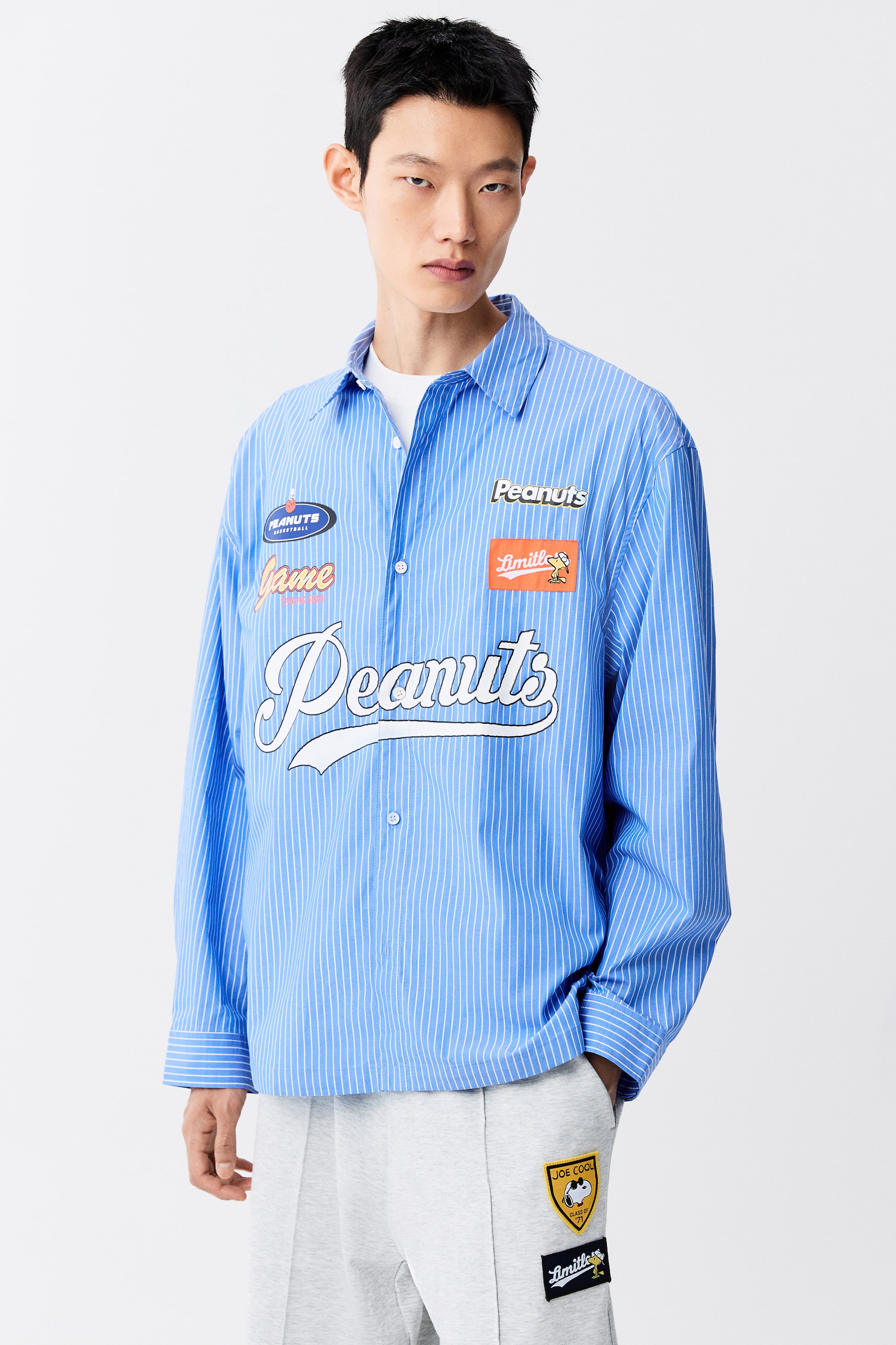 View larger image: The model is wearing a blue-and-white vertically striped long-sleeved button-up shirt featuring several cartoon-themed graphic patches and a large white script logo across the front. The shirt is layered over a plain white T-shirt and styled with light grey sweatpants.
