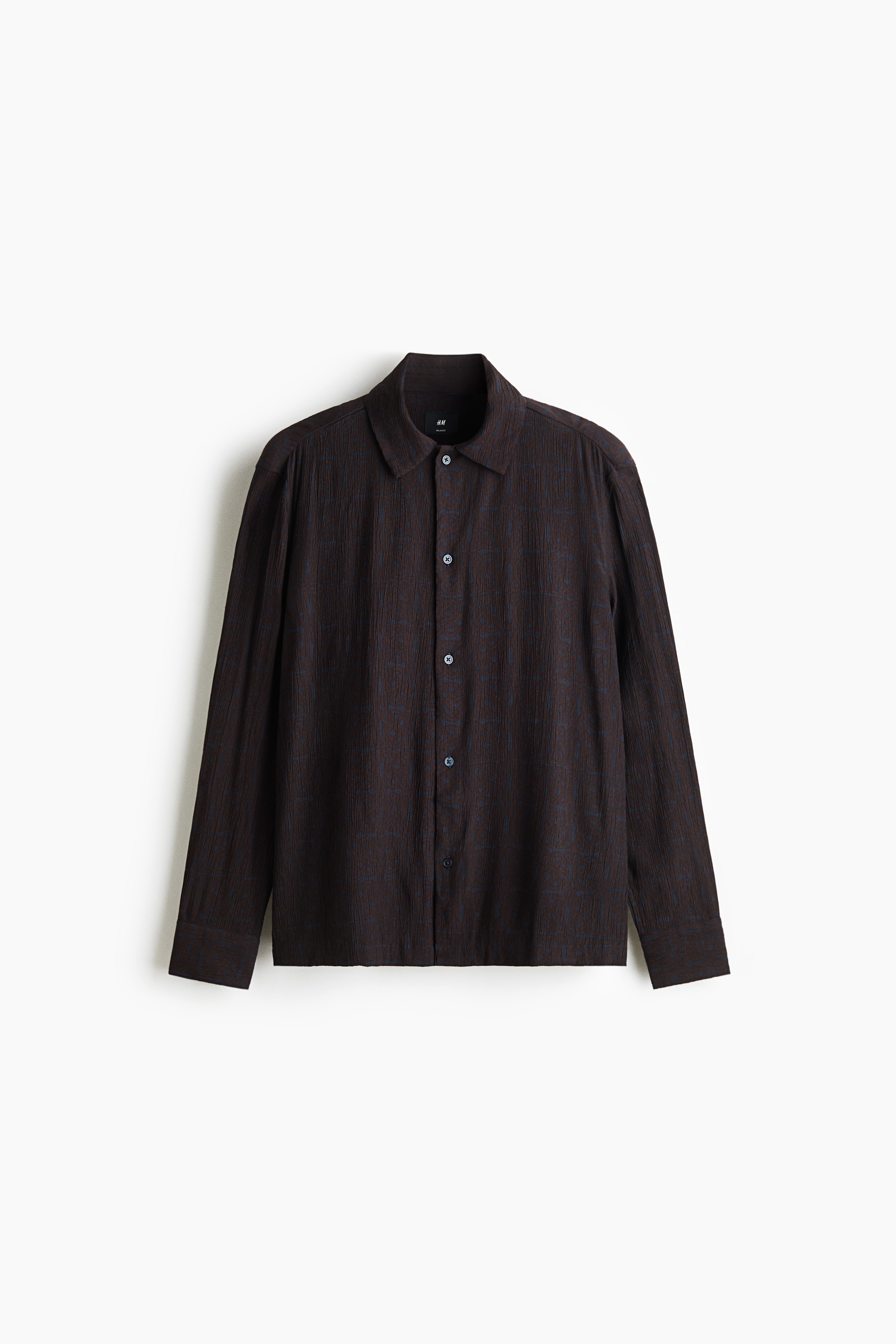 The model is wearing a dark brown shirt in a textured viscose blend, featuring a relaxed fit, a collar, and a button-down front. The long-sleeved shirt is styled with a partial tuck into dark blue jeans, completed with a brown leather belt.