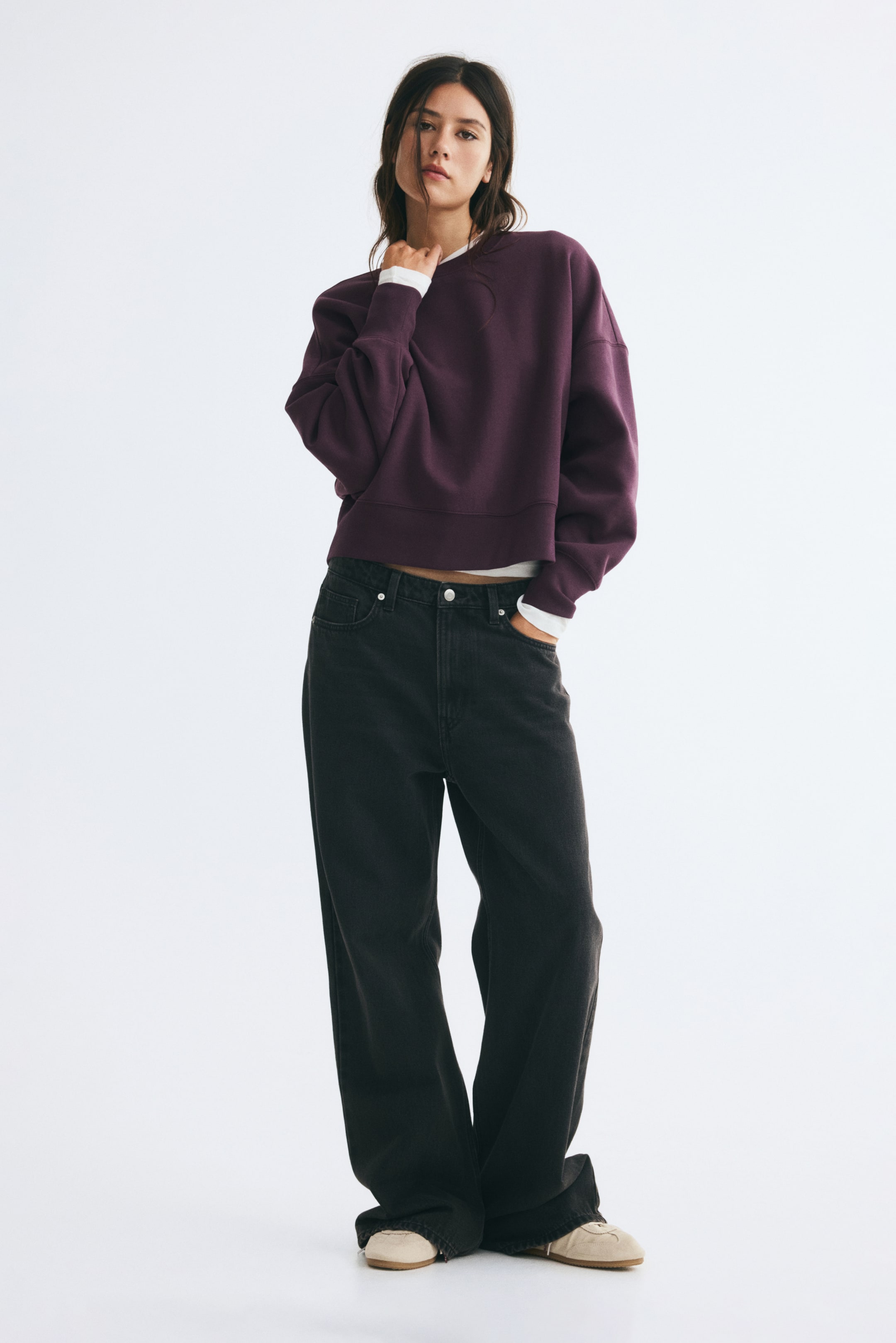 Büyük resmi görüntüle: Modelin üzerinde, yuvarlak yaka, düşük omuzlar ve manşetlerde ile etek ucunda ribanalı bitiş detaylarına sahip, büyük beden koyu mor bir sweatshirt yer alıyor. Sweatshirt, beyaz uzun kollu bir üst katman üzerine giyilmiş ve siyah geniş paça jeanlerle kombinlenmiş; modelin bir eli yaka kısmına yakın şekilde duruyor.
