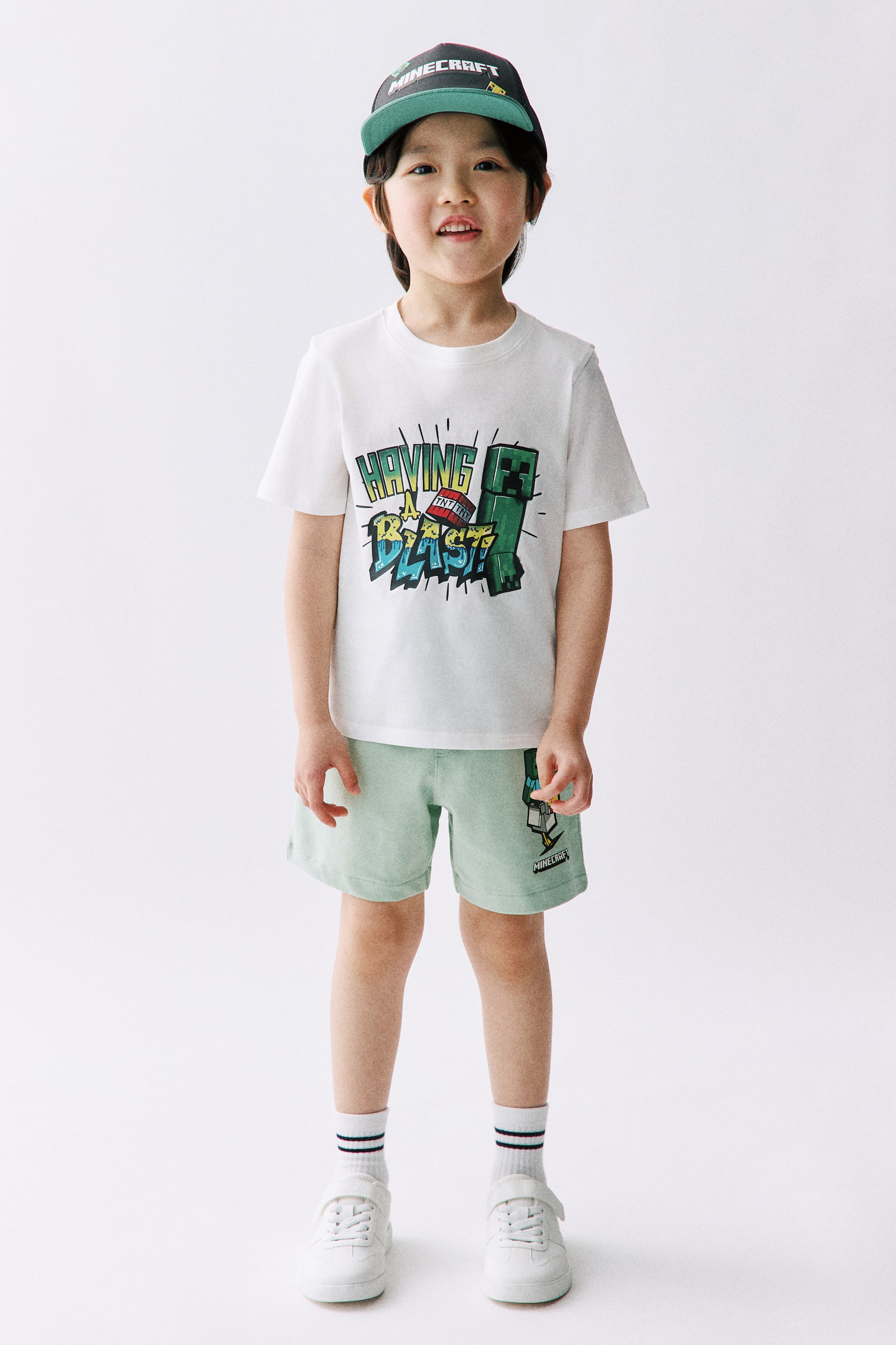 拡大画像を表示: 子供用の服装は、白い半袖Tシャツに「HAVING A BLAST!」というグラフィックがあり、緑のマインクラフトのクリーパーと赤いTNTブロックが描かれています。この服装には、左足に小さなツルハシと「MINECRAFT」のグラフィックが描かれたライトグリーンのショーツ、前面に「MINECRAFT」と刺繍された緑のつば付きのダークキャップ、白いストライプのアンクルソックスの上に履くフックアンドループファスナー付きの白いスニーカーも含まれています。