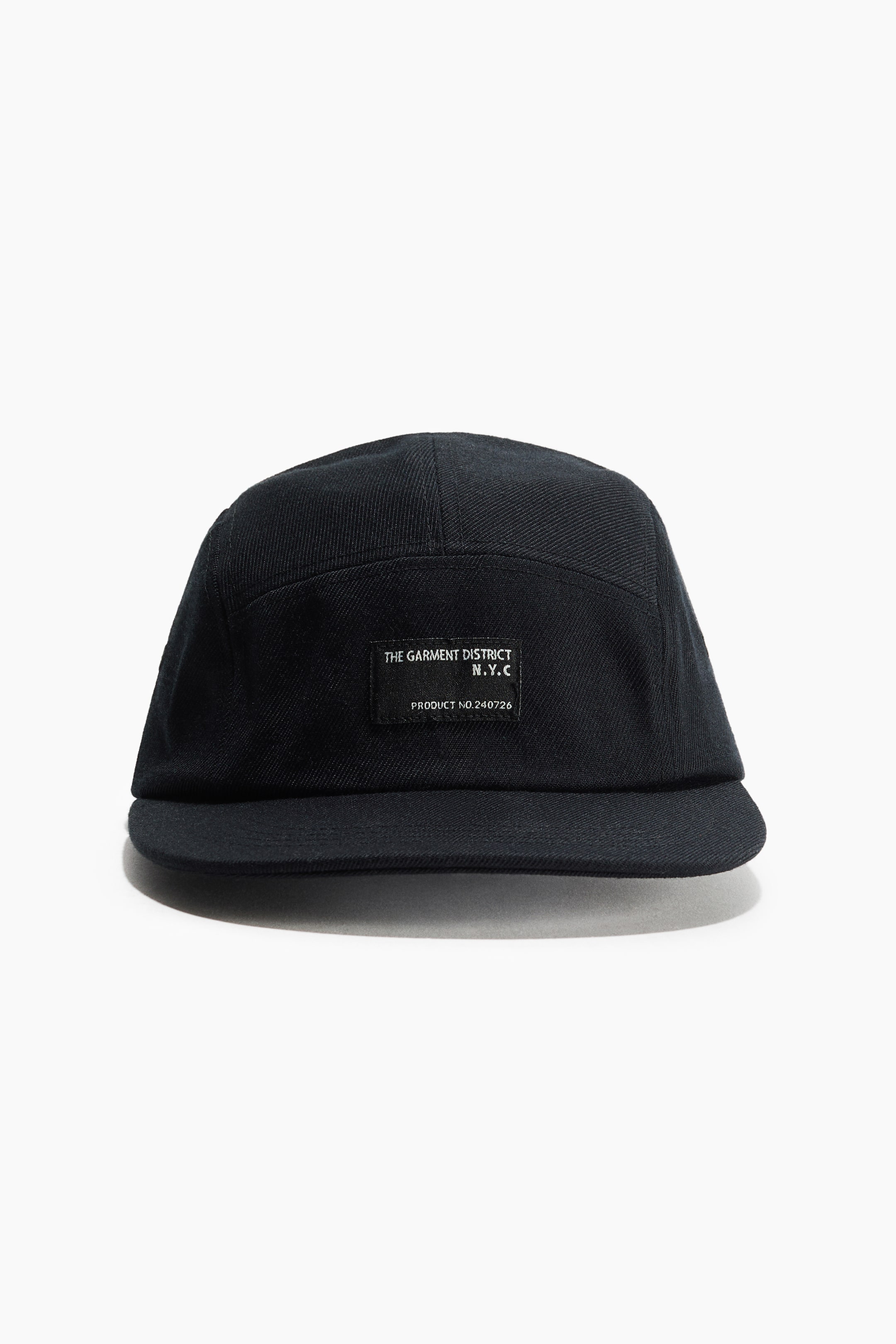 View larger image: A black 5-panel cap is angled slightly to the right, featuring a woven texture and a flat brim. A rectangular black label, displaying white text that reads "THE GARMENT DISTRICT N.Y.C PRODUCT NO. 240726", is affixed to the front panel.