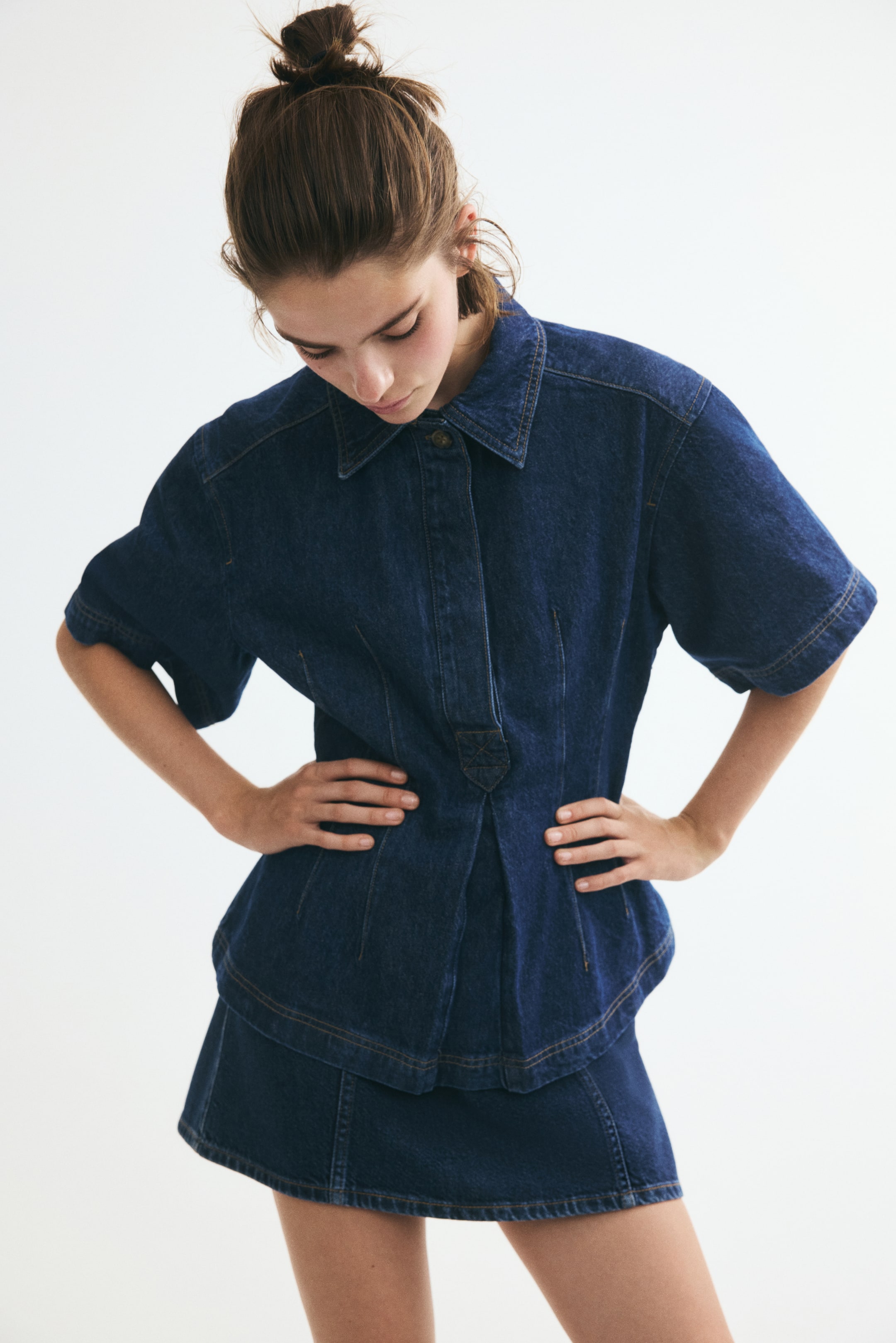 View larger image: A short-sleeved shirt in rigid cotton denim, featuring a collar with a visible button on the collar stand, a concealed button placket, an inverted box pleat at the front hem, and shaping waist darts for a nipped-in silhouette, is styled on the model with a matching denim skirt. The model is posing with hands on hips.