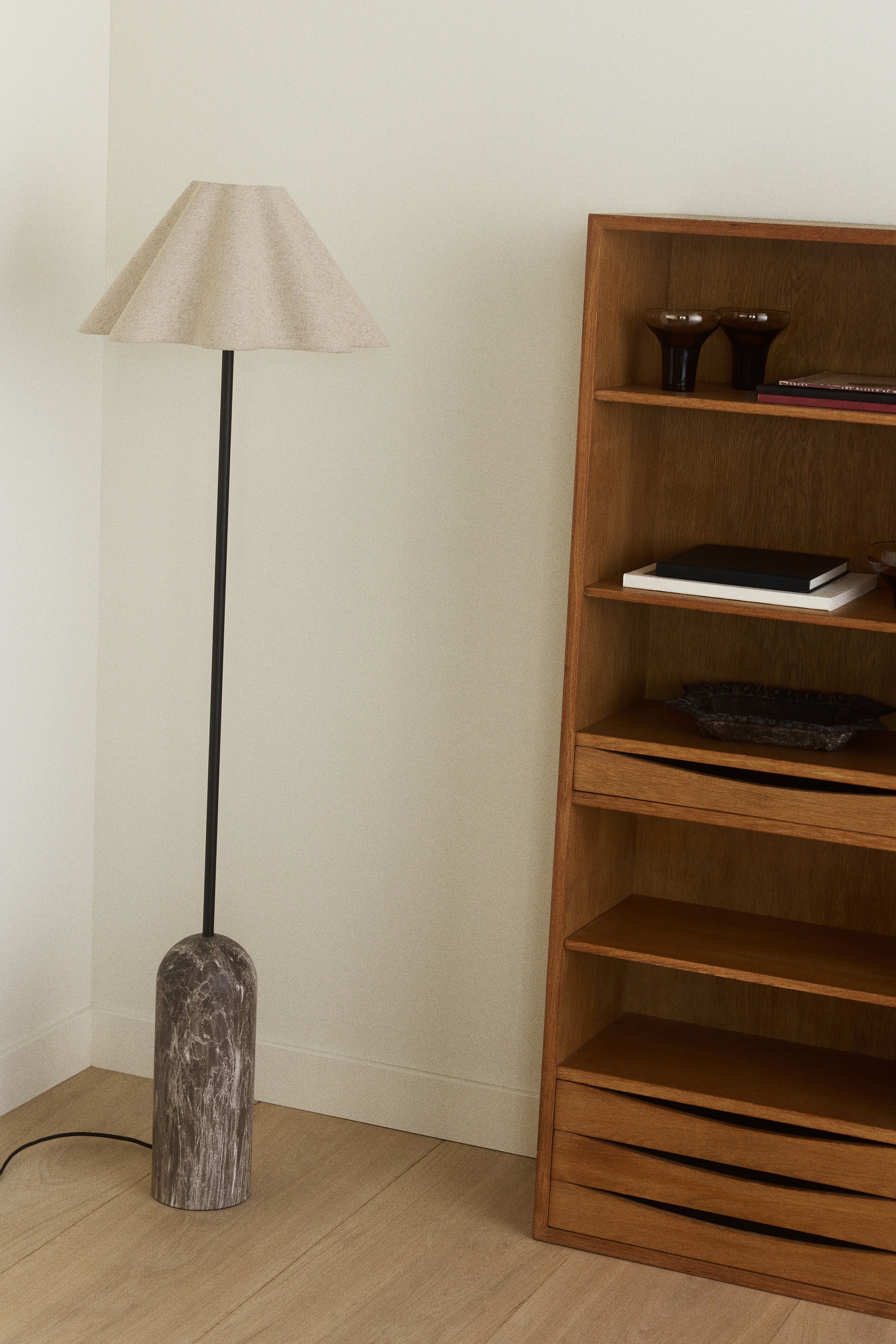 Een lamp staat rechtop, voorzien van een lichtbruine, geplooide kegelvormige kap. Een dunne zwarte steel verbindt de kap met een donkerbruine, cilindrische marmeren voet met afgeronde randen, waaruit een zwart snoer zich uitstrekt.