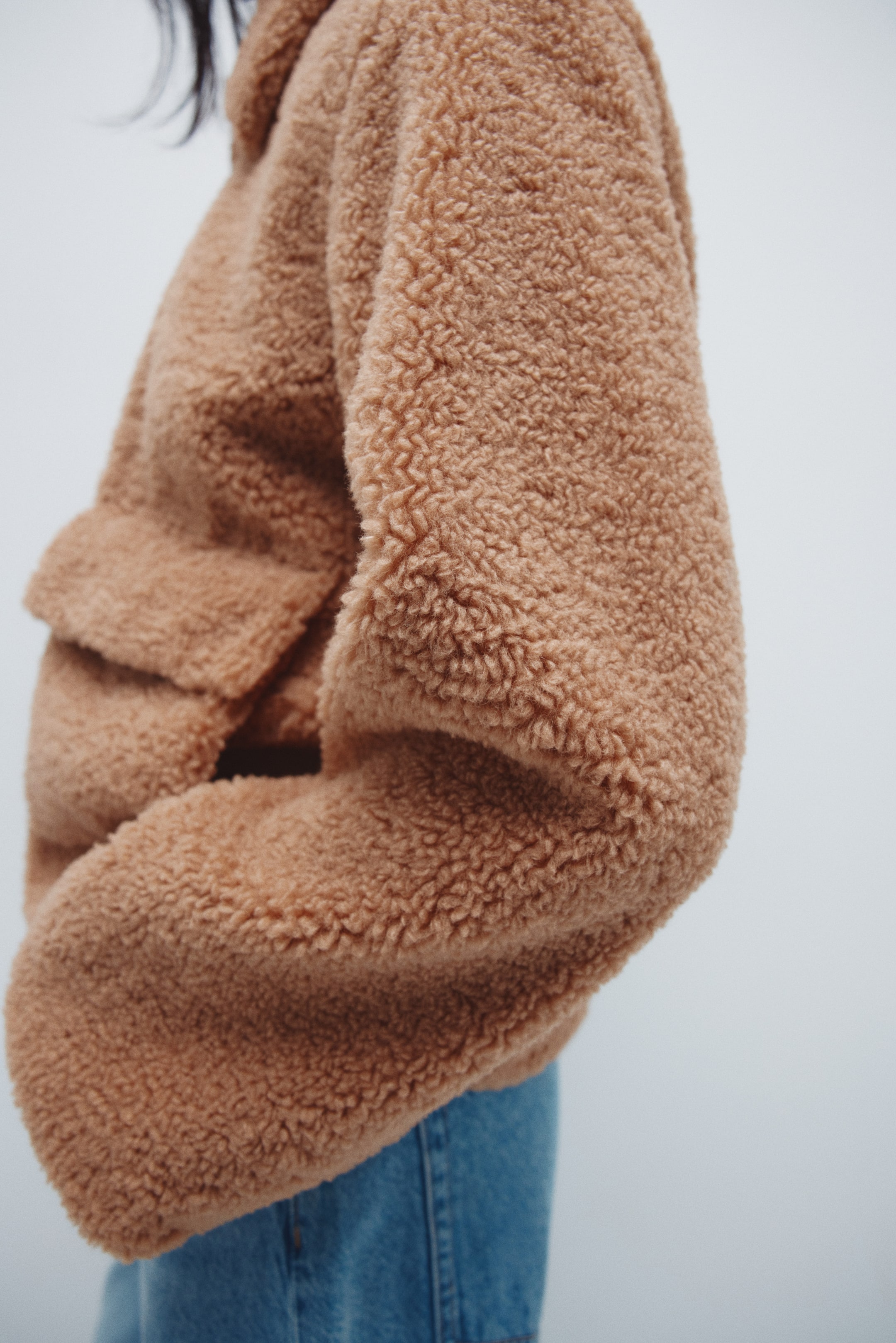 Voir une image plus grande: Une veste en polaire à effet nounours couleur fauve, avec un tissu à texture bouclée et une poche à rabat visible, est montrée de biais en angle de côté avant, associée à un jean bleu.