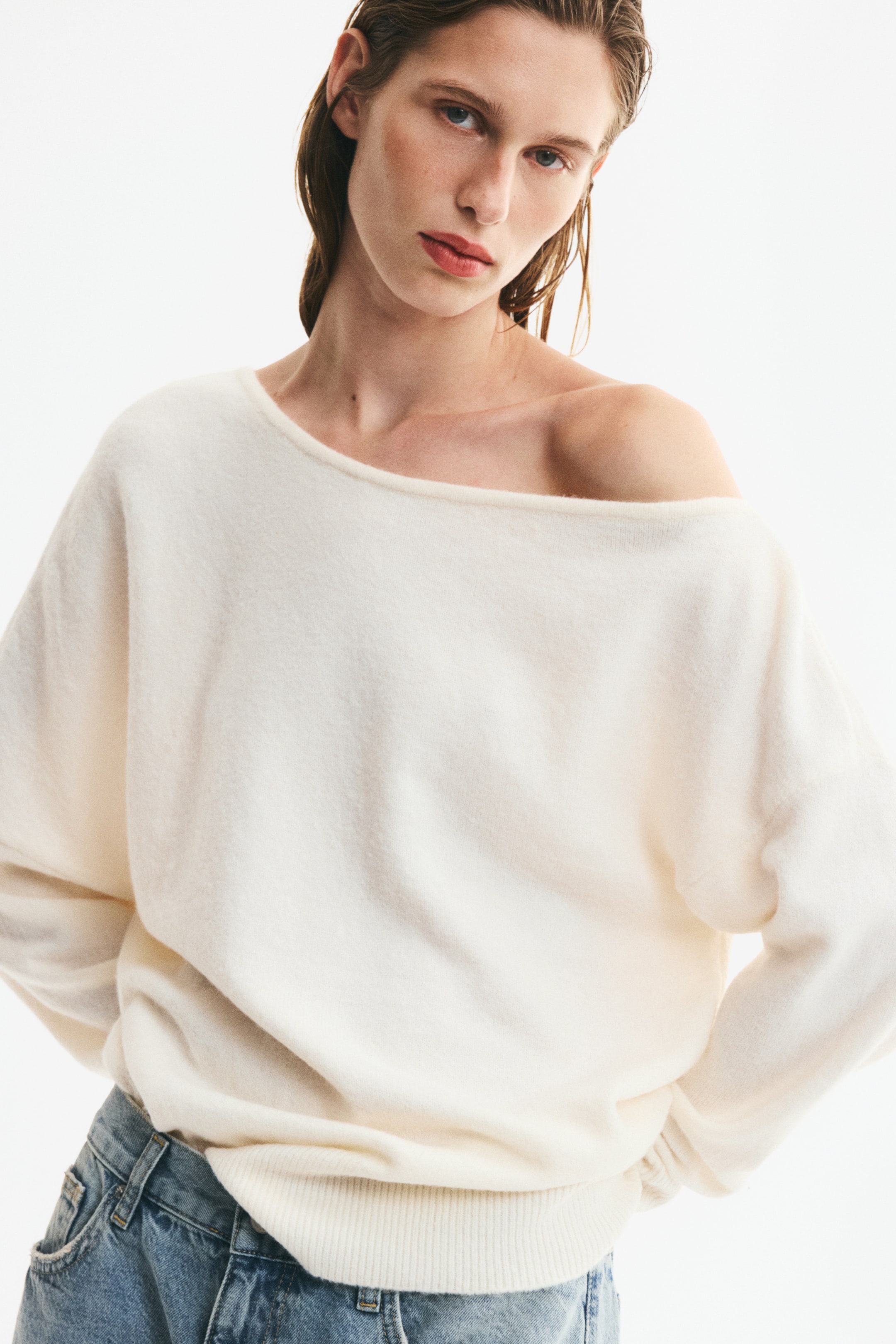 View larger image: An off-white, fine-knit sweater featuring an asymmetrical, one-shoulder neckline and dropped shoulders is being worn by the model. The long-sleeved sweater has a ribbed hem and is styled tucked into blue denim jeans.