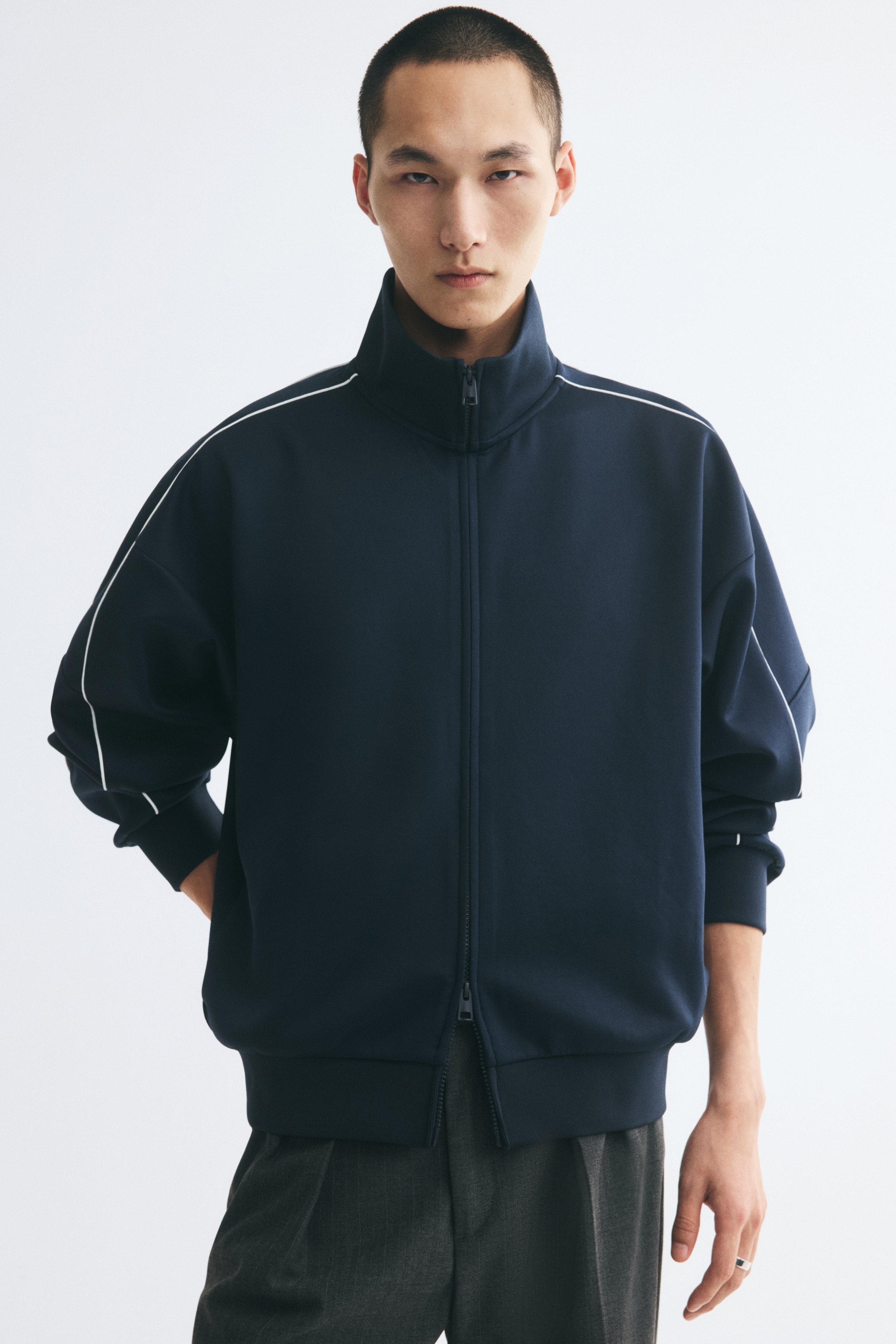 Má»t chiáº¿c Ã¡o khoÃ¡c thá» thao zip mÃ u xanh navy Äang ÄÆ°á»£c chá»¥p chÃ­nh diá»n, ná»i báº­t vá»i cá» Äá»©ng cao vÃ  phom dÃ¡ng thoáº£i mÃ¡i, vai trá». ÄÆ°á»ng viá»n tráº¯ng máº£nh cháº¡y dá»c theo pháº§n vai vÃ  xuá»ng tay Ã¡o, káº¿t thÃºc báº±ng bo tay Ã¡o vÃ  gáº¥u Ã¡o bo gÃ¢n Äá»ng mÃ u.
