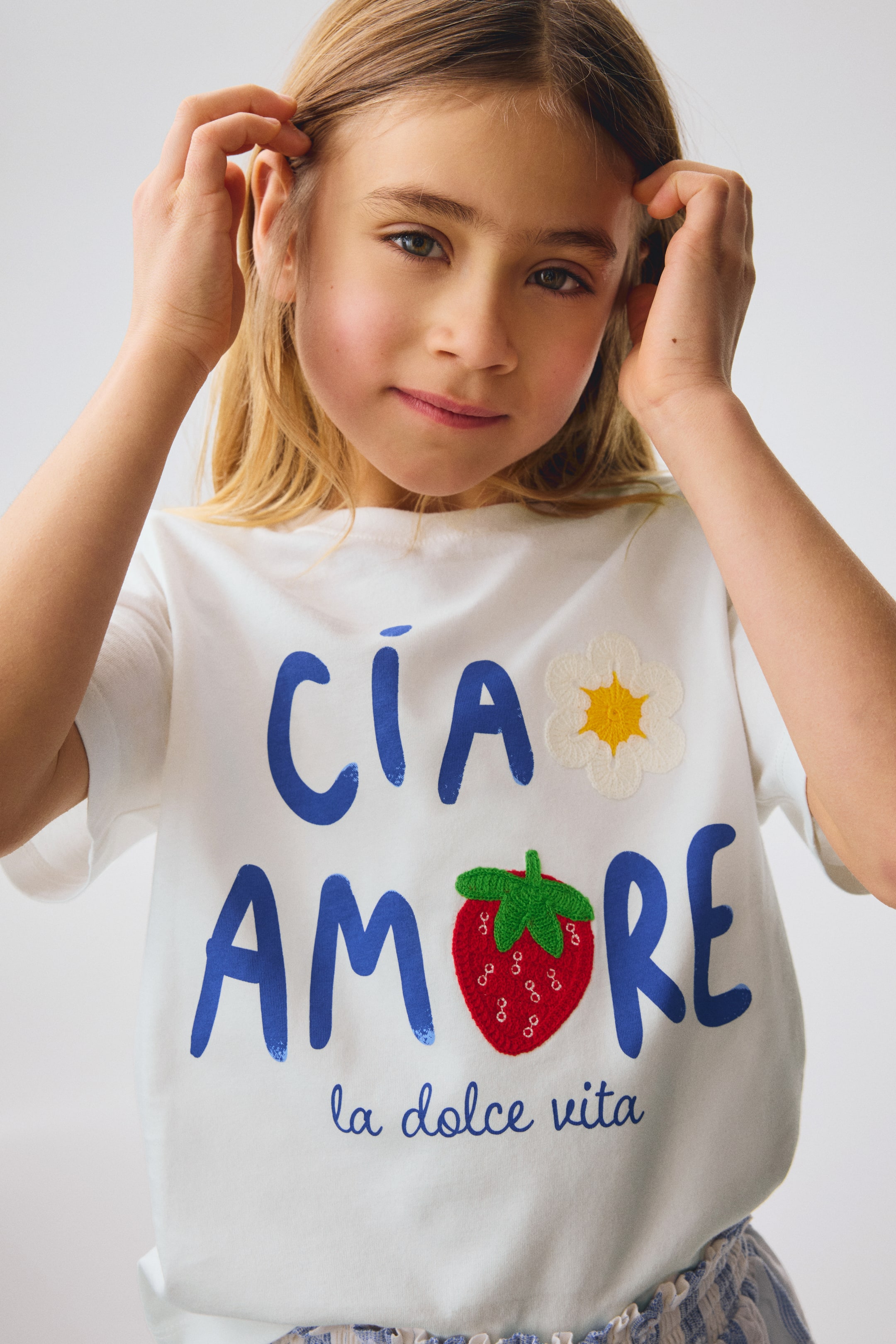 Xem ảnh lớn hơn: Một chiếc áo thun trắng có hình in lớn màu xanh "CIA AMORE" ở phía trước, được nhấn nhá bằng một bông hoa trắng đính nổi với nhụy vàng gần chữ "CIA". Một quả dâu tây đỏ có kết cấu với lá xanh cũng được đặt trong chữ "AMORE", với dòng chữ "la dolce vita" viết nhỏ màu xanh phía dưới.
